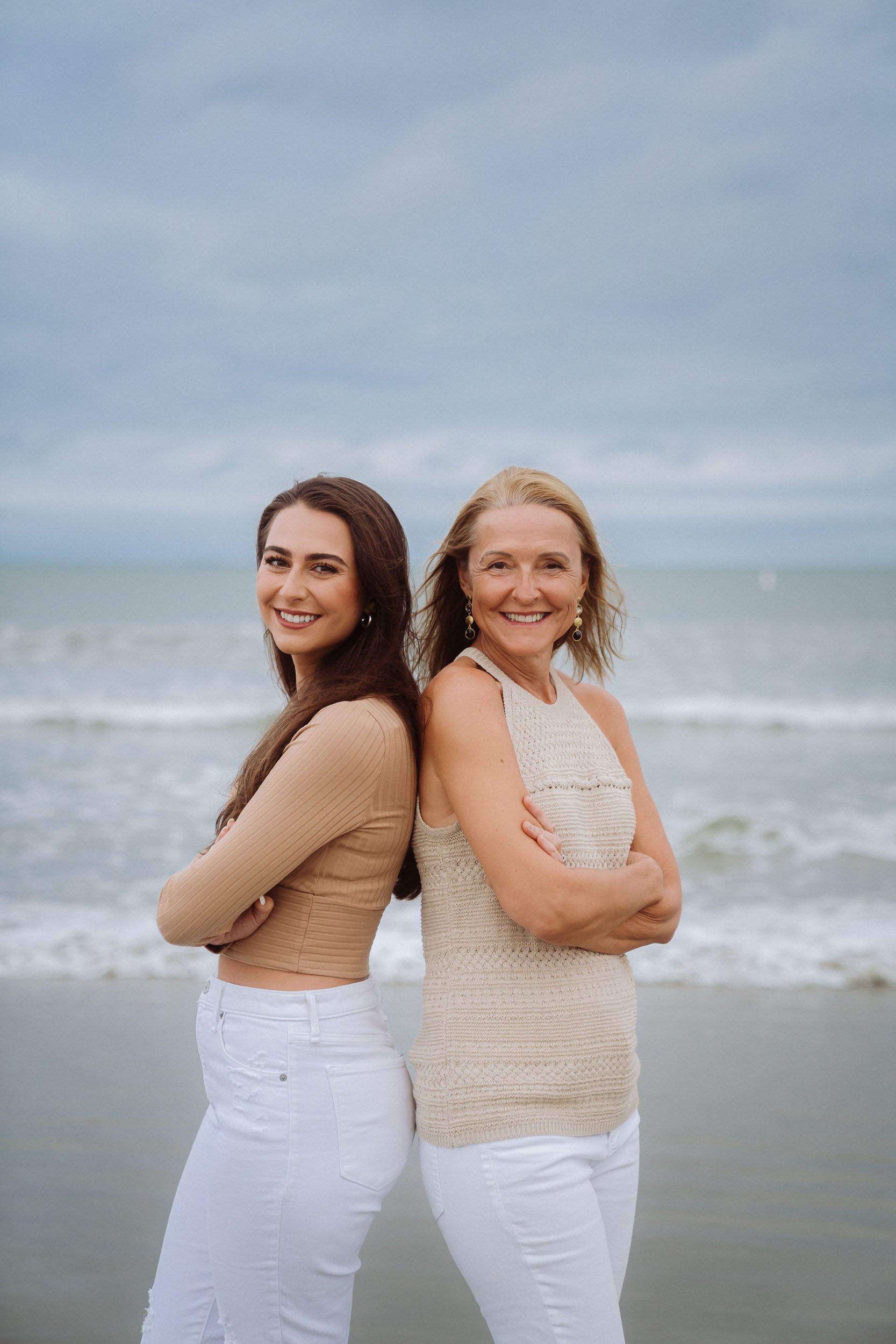 Two women standing back to back on a beach with arms crossed, smiling at the camera. Overcast sky and ocean in the background.