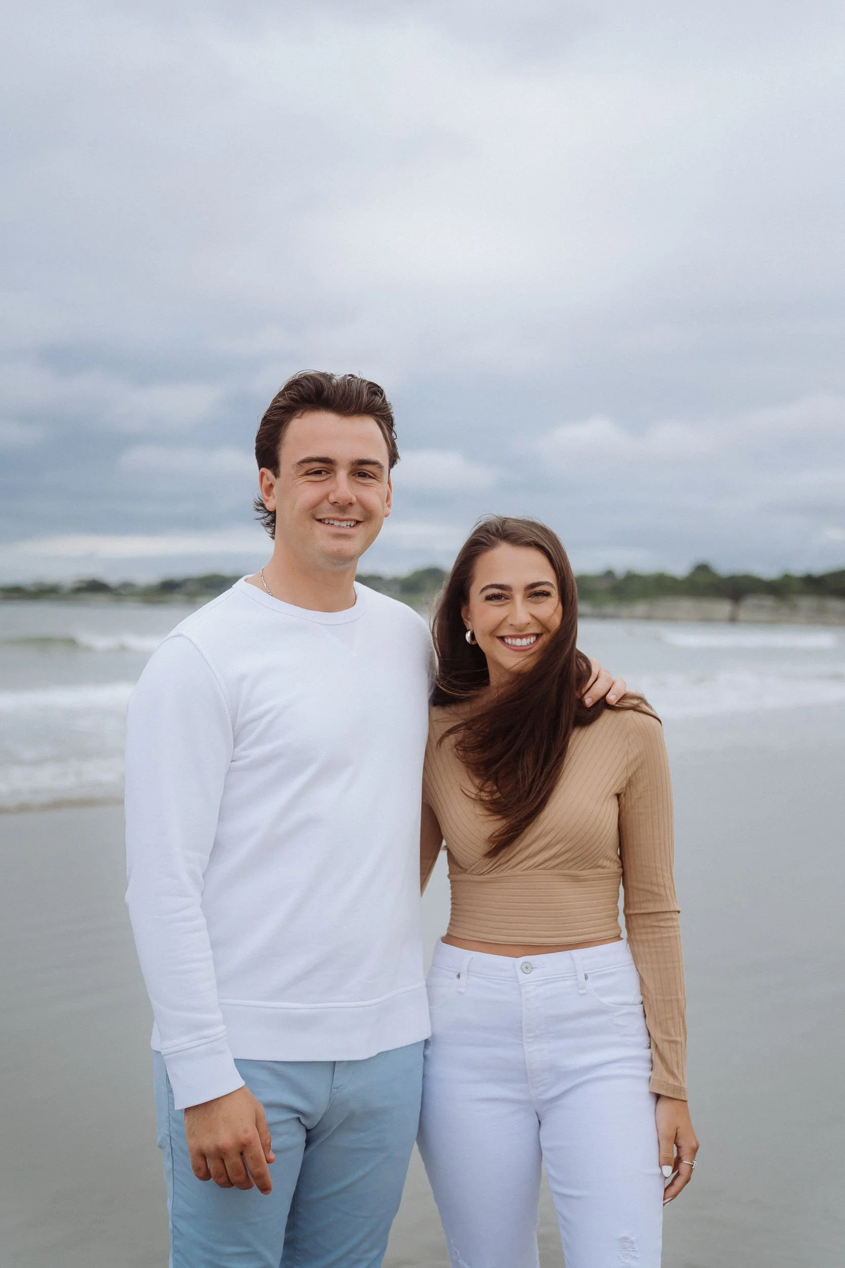 A smiling young man and woman standing on the beach, close to each other, with the ocean and cloudy sky in the background.