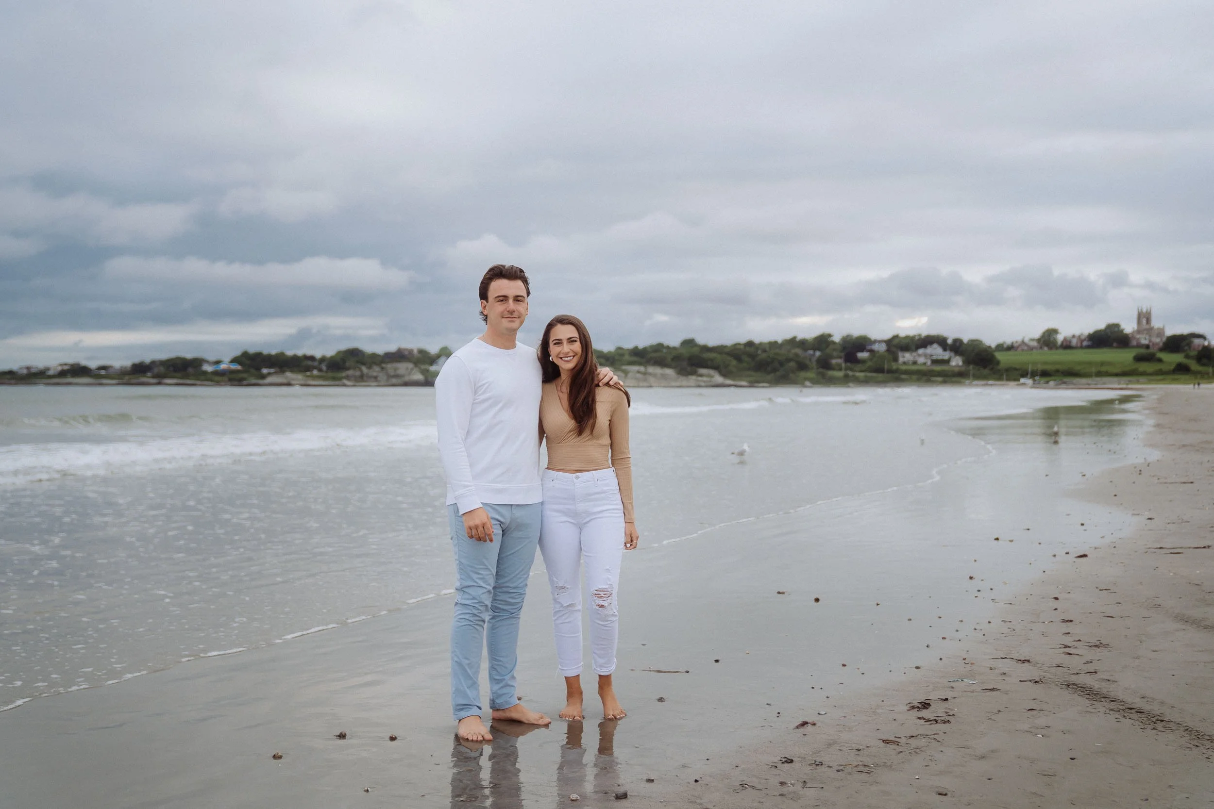 A young man and woman standing close together on a beach, smiling, with arms around each other. The woman is dressed in a beige long-sleeve top and white ripped jeans, the man in a white long-sleeve shirt and light blue pants. The beach has wet sand,