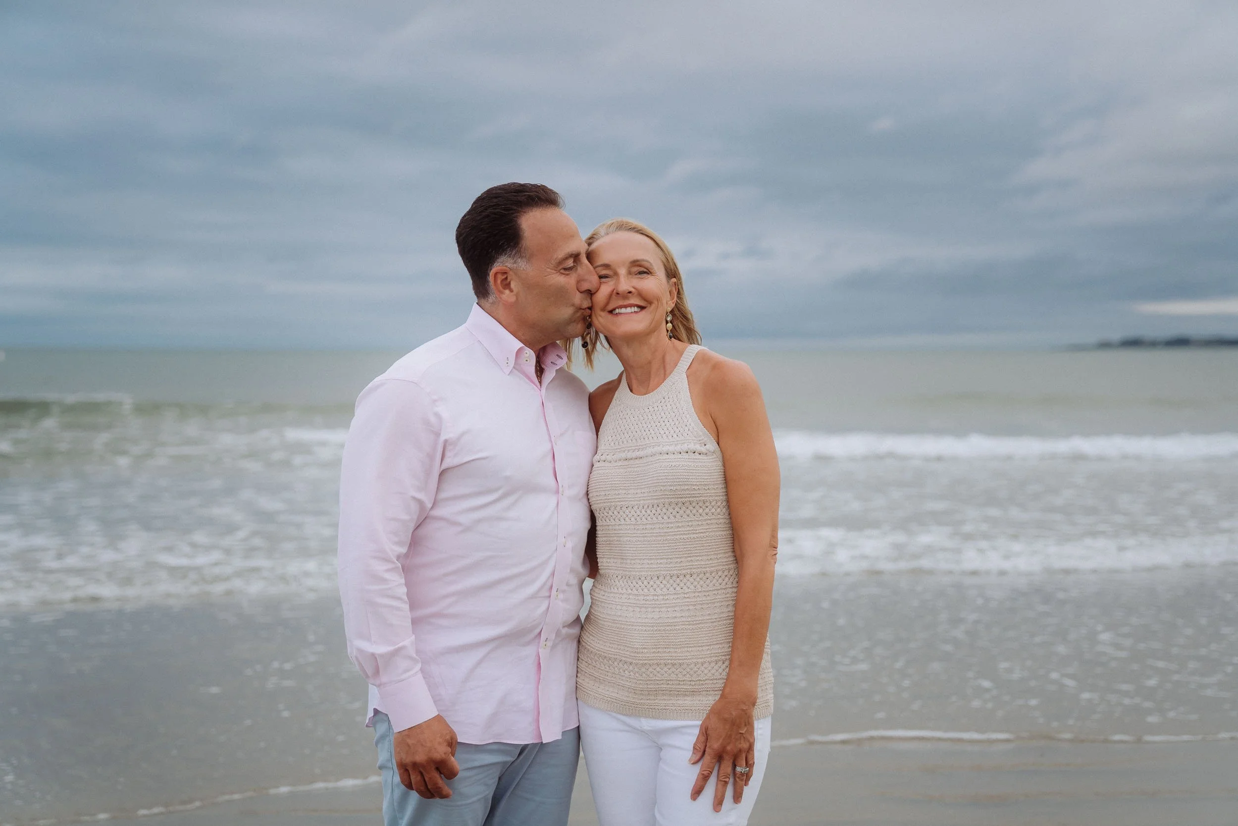 A middle-aged man kisses a smiling middle-aged woman on the cheek at the beach, with waves and a cloudy sky in the background.