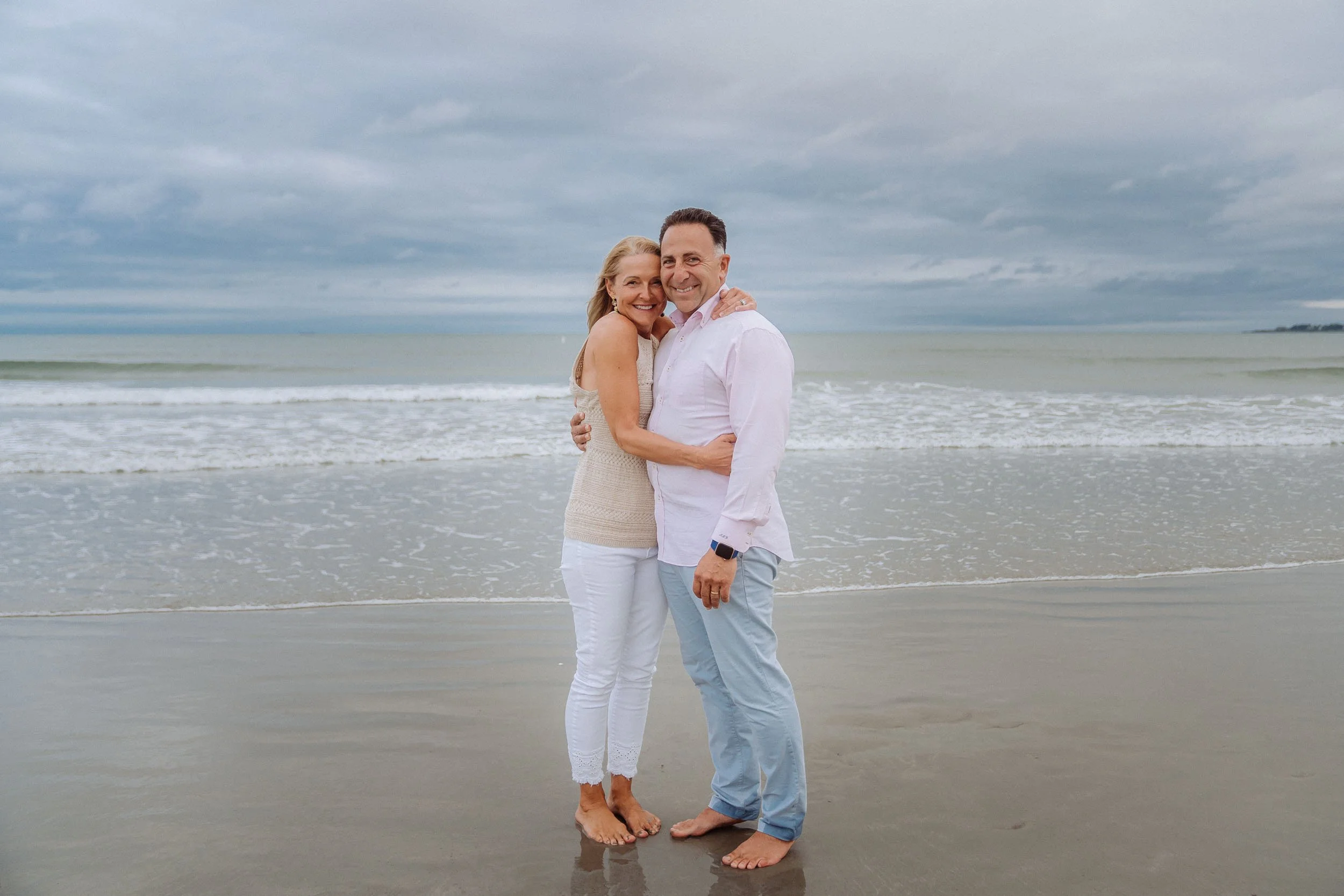 A happy couple hugging on a beach with cloudy skies.