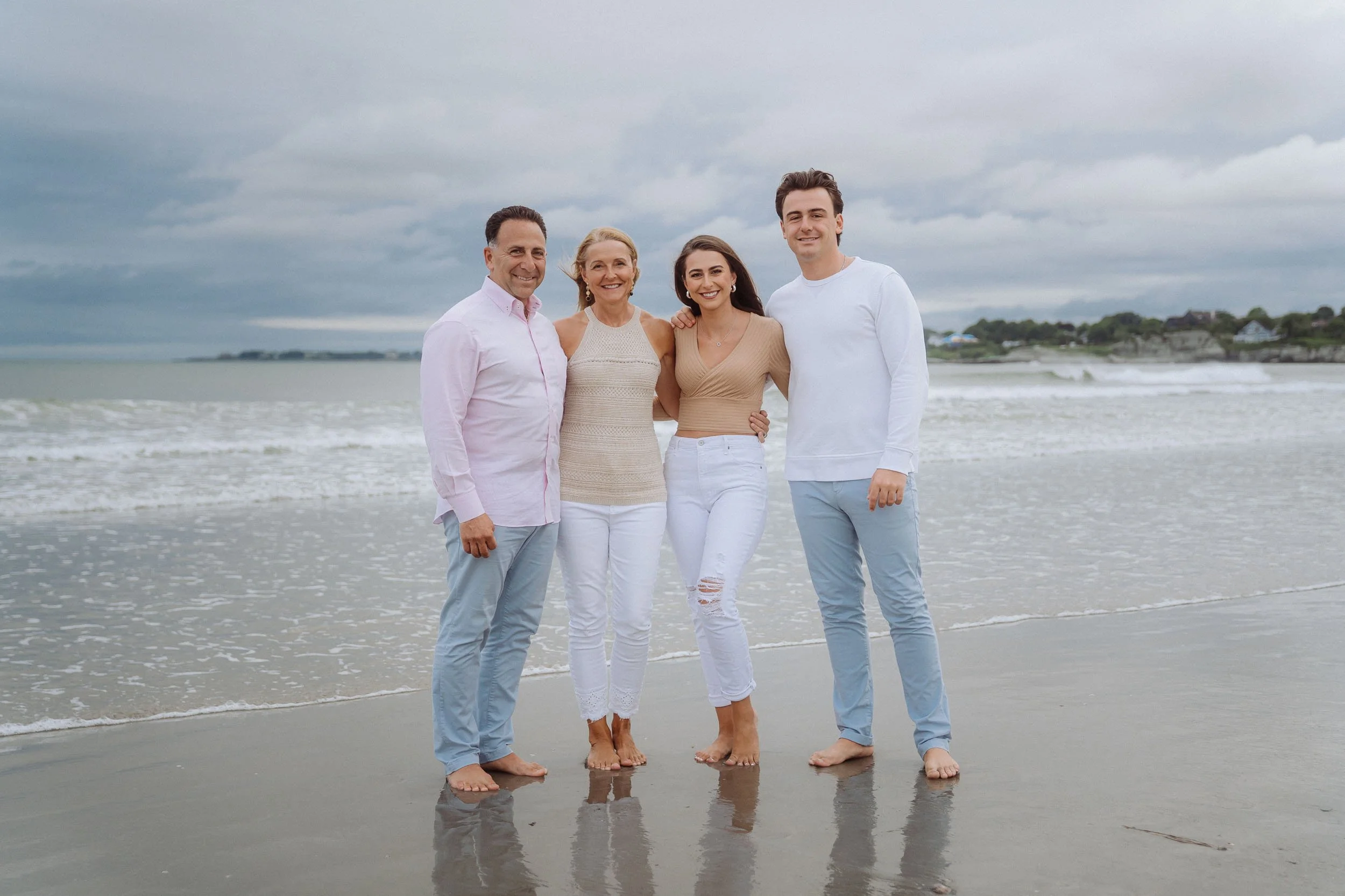 A family of four standing on a sandy beach in the rain, smiling and posing for a photo with waves in the background.