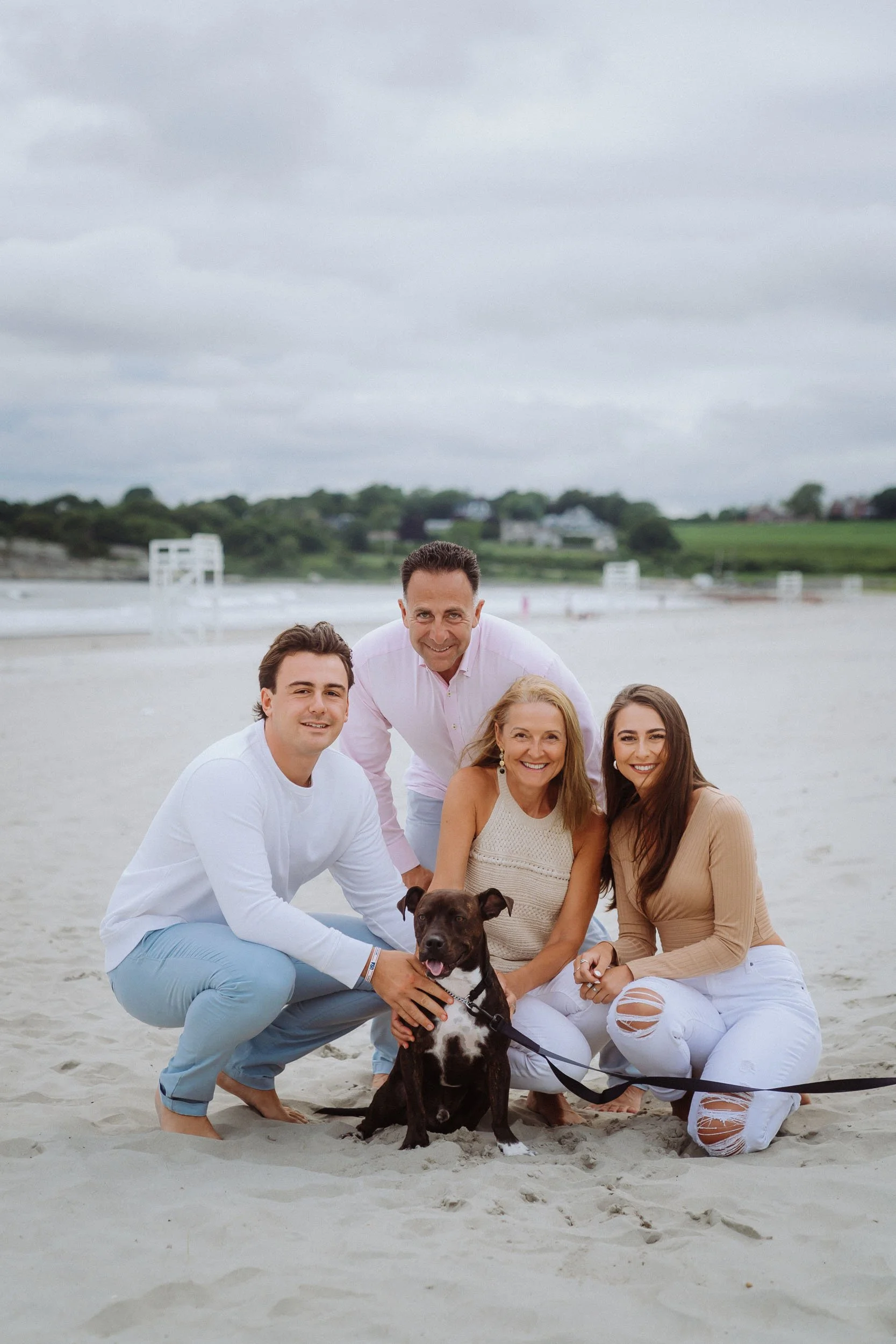 Group of five people and a dog at the beach, smiling and posing for the photo on sand with dunes and cloudy sky in the background.
