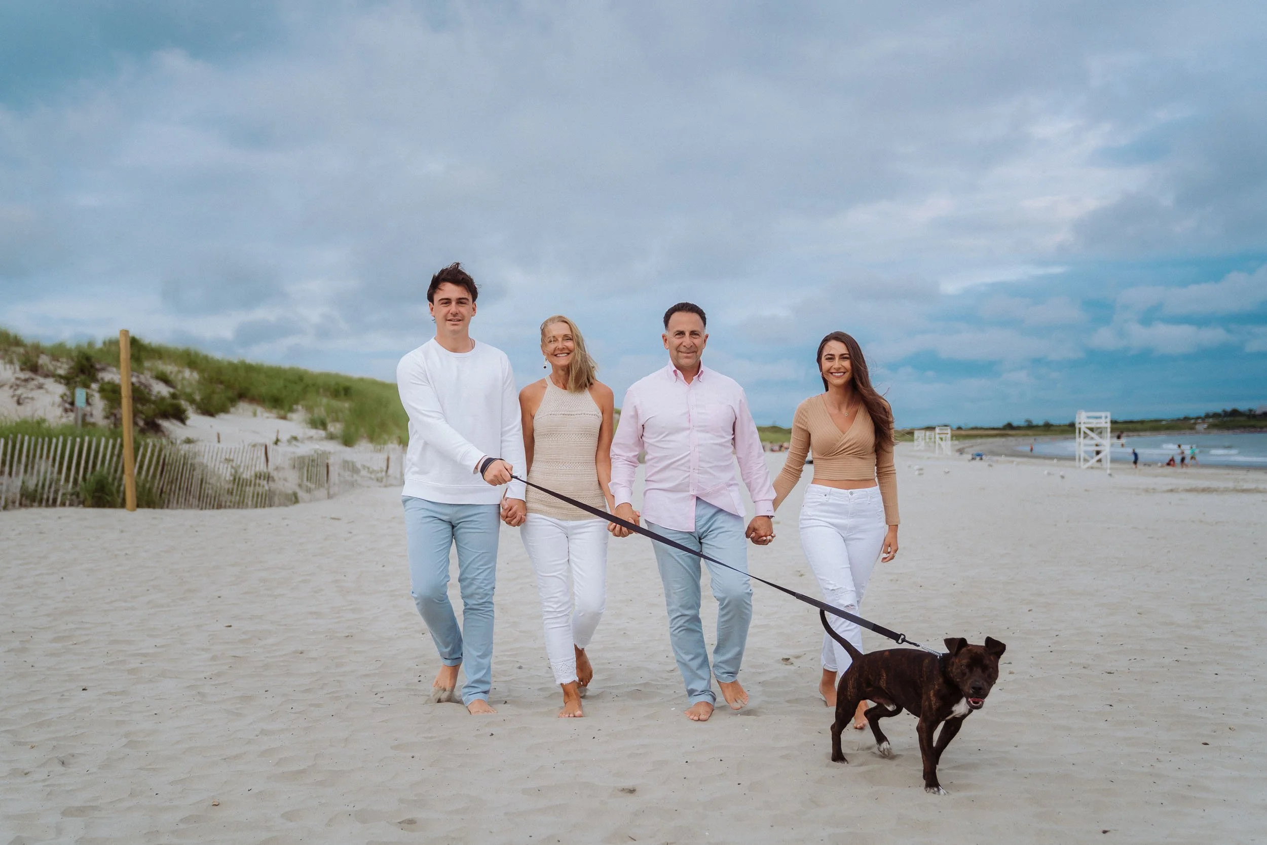 A family walking on a sandy beach holding hands, with a dog on a leash, under a cloudy sky.