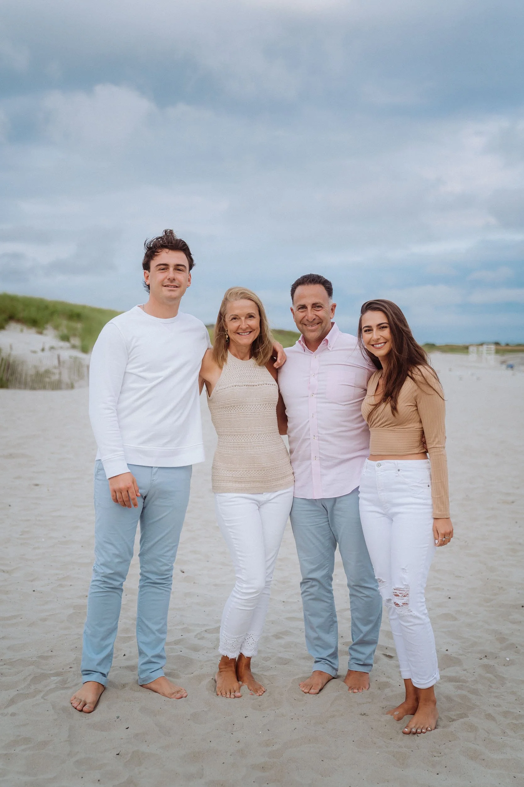 A family of four standing on a sandy beach with a cloudy sky in the background. They are all smiling and dressed in light-colored casual clothing.