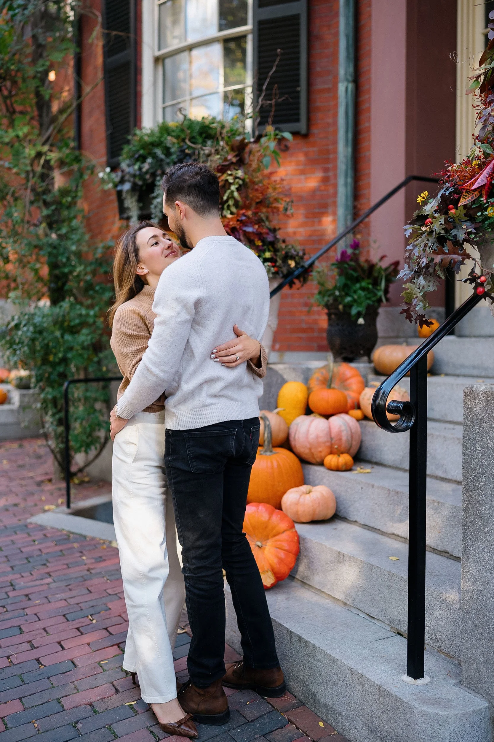 A couple embraces on a brick sidewalk in front of a house decorated with pumpkins and fall foliage.