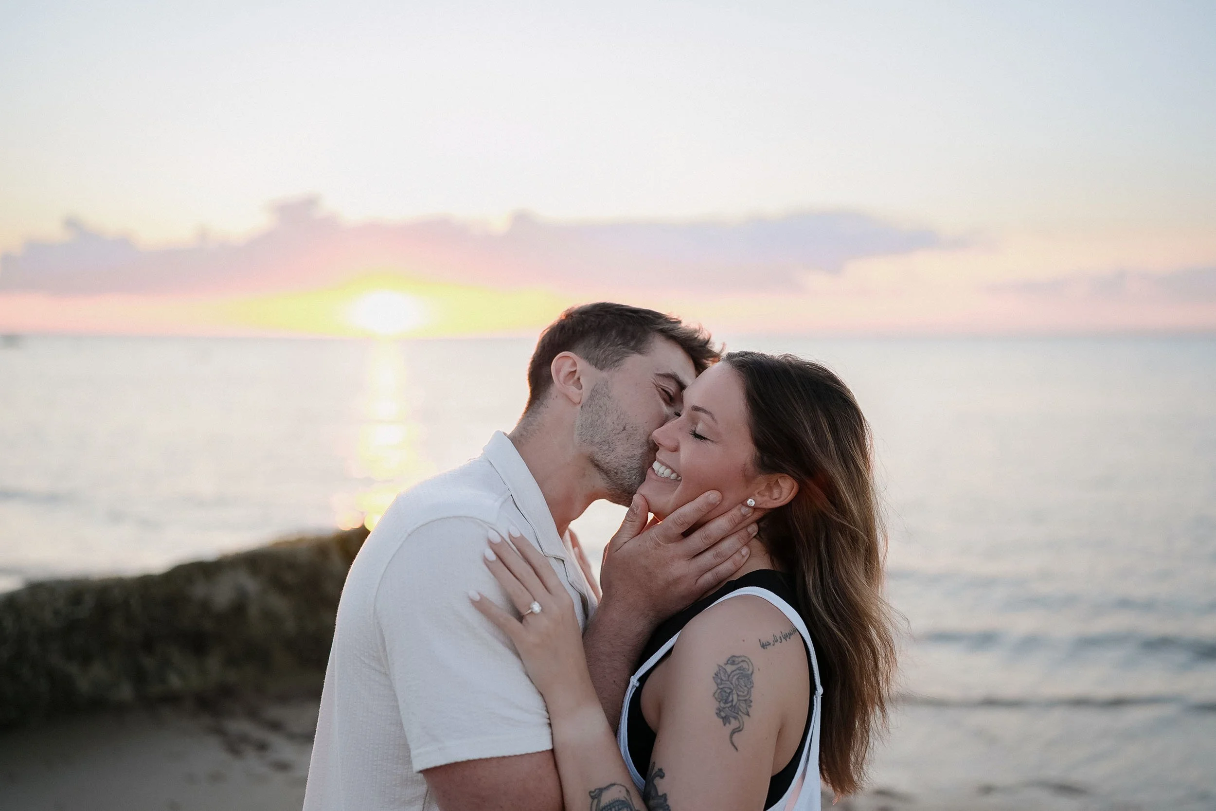 A young couple hugging and about to kiss on a beach during sunset, with the ocean and a rocky shoreline in the background.