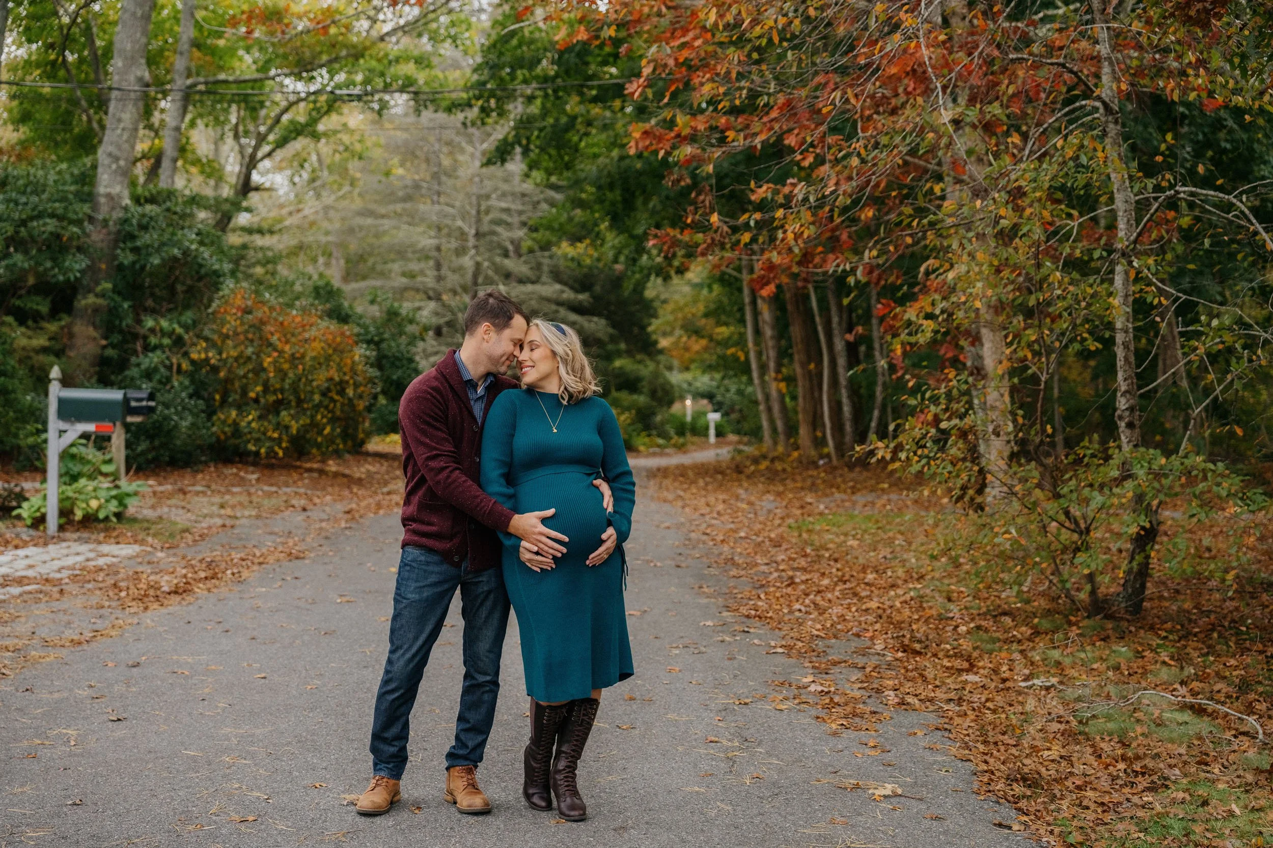A pregnant woman and a man standing close together on a tree-lined street with autumn leaves, smiling and touching her belly.