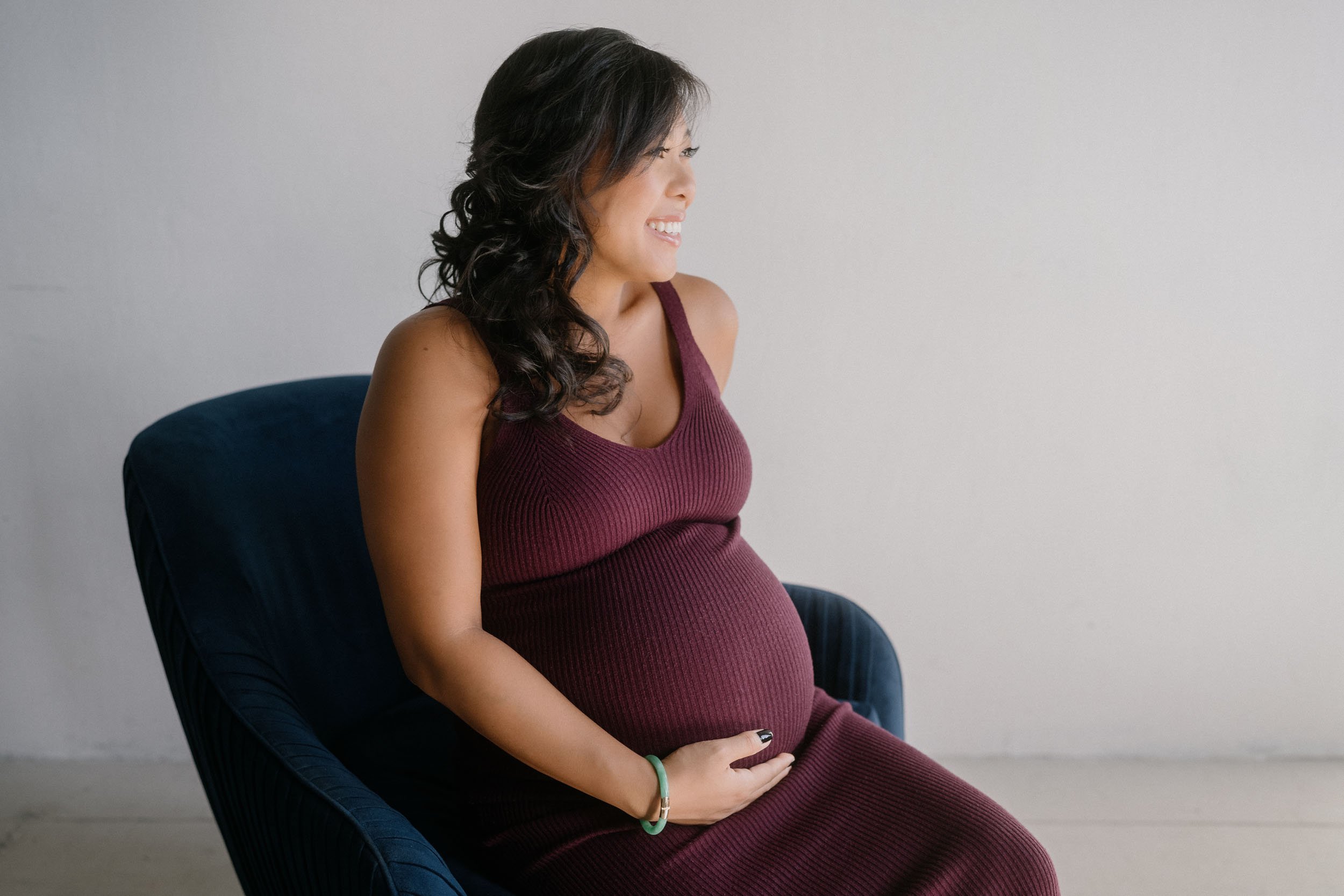 A pregnant woman with dark, curly hair sitting on a navy blue chair, smiling and looking to her right, wearing a sleeveless maroon dress.
