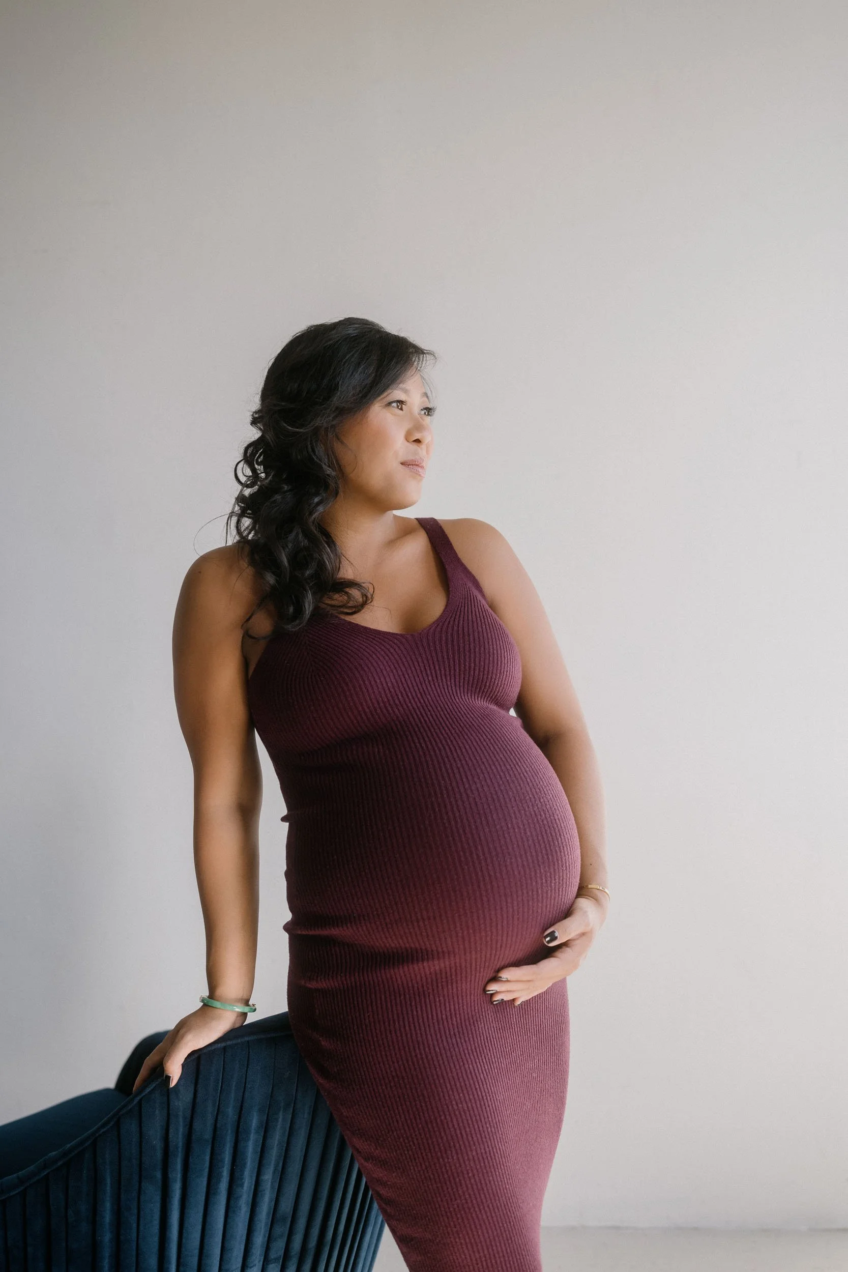 Pregnant woman in a burgundy sleeveless dress standing near a dark blue velvet chair, looking to her left, with her right hand resting on the chair and her left hand on her belly, against a plain light background.