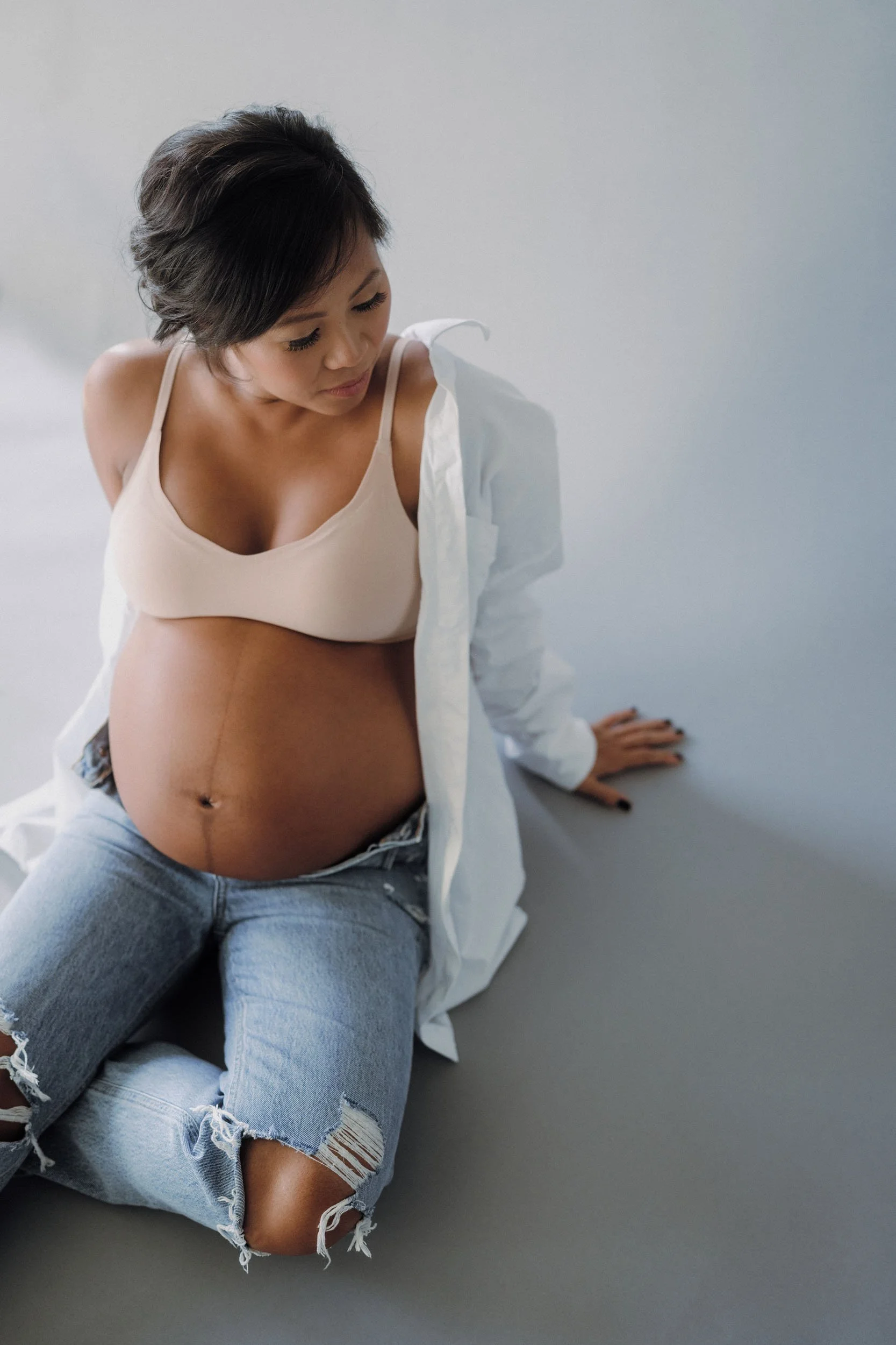 A pregnant woman sitting on the floor, wearing a beige bra, a white shirt, and ripped jeans, looking down.