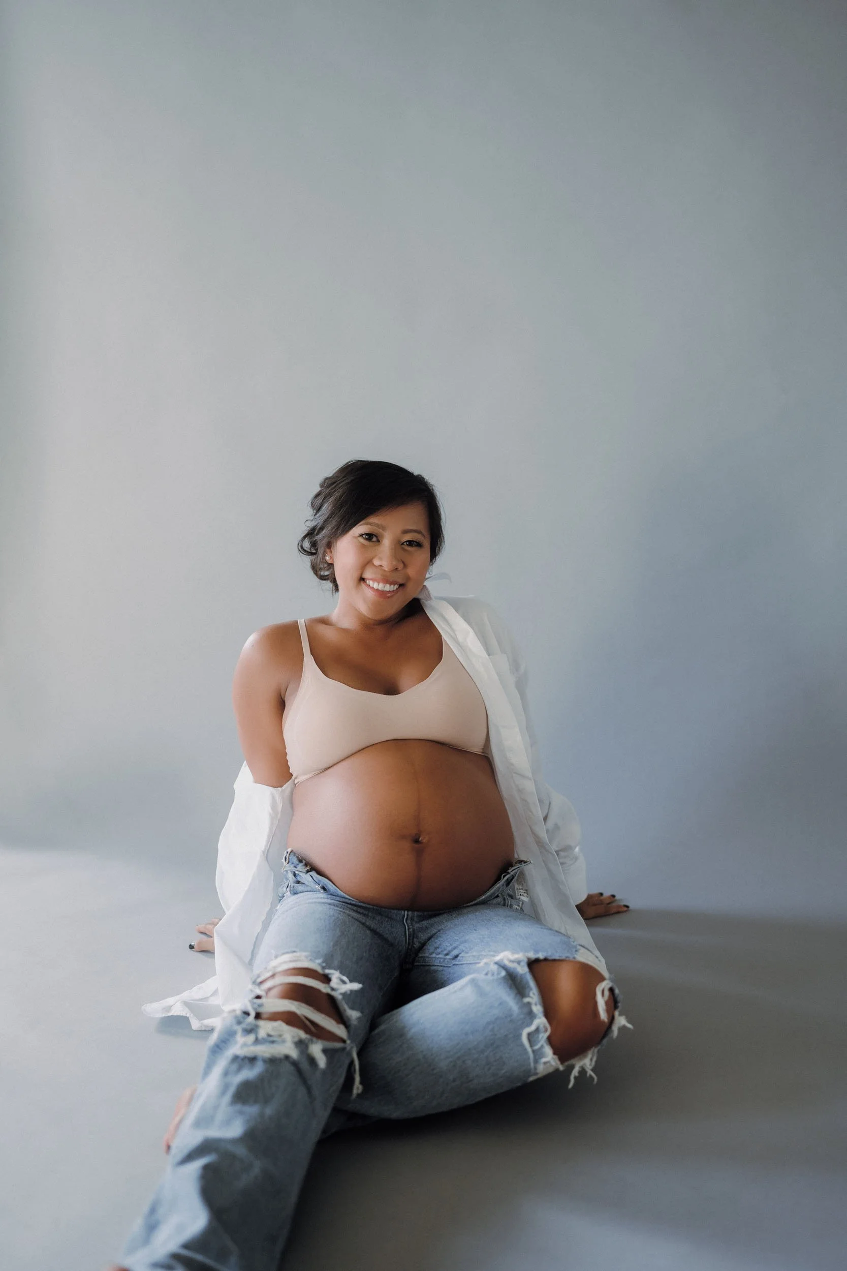 A pregnant woman smiling while sitting on the floor in front of a plain background, wearing a beige tank top, ripped jeans, and an open white shirt.