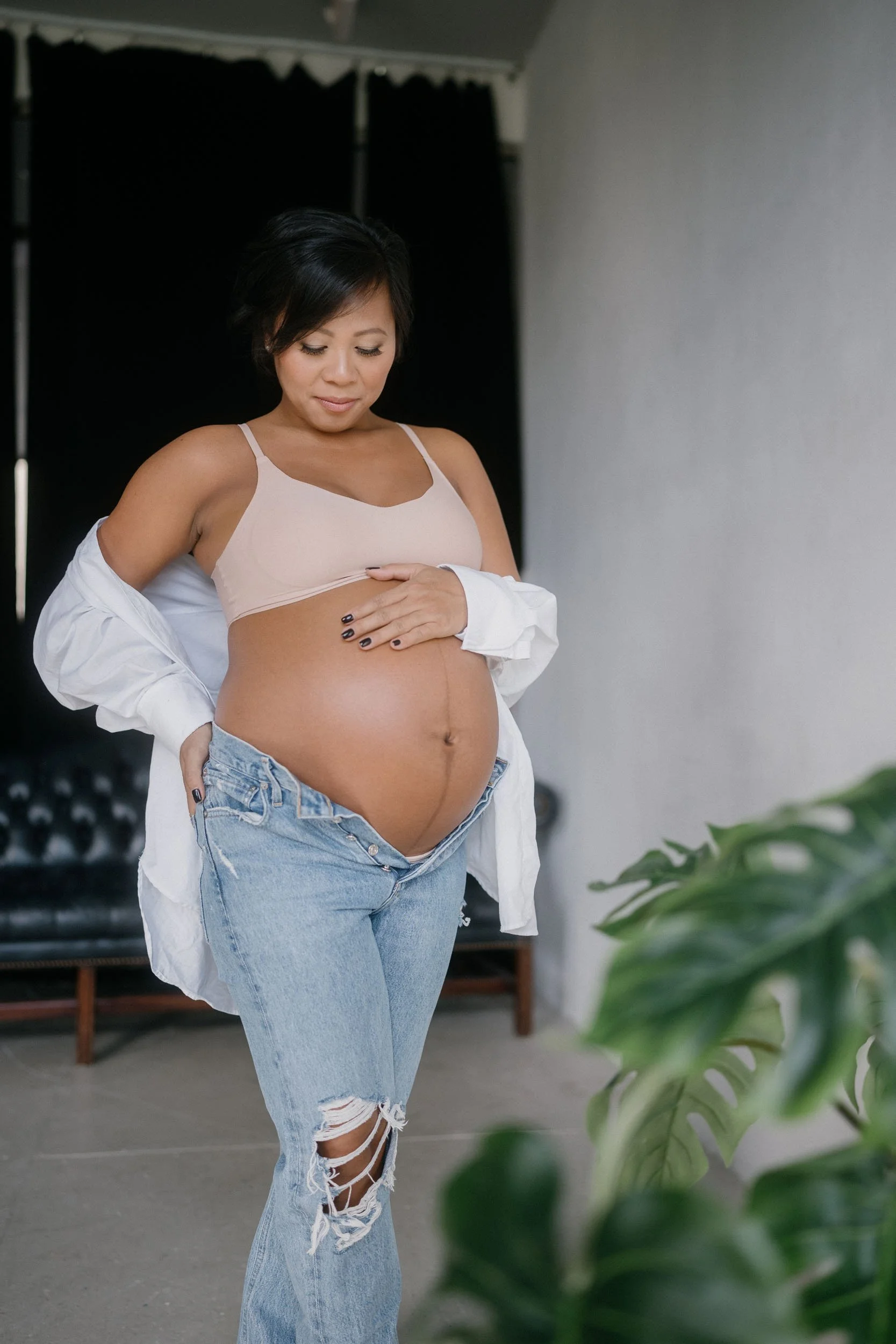 A pregnant woman standing indoors, wearing a beige crop top, ripped jeans, and an unbuttoned white shirt, gently holding her baby bump and looking down at it.