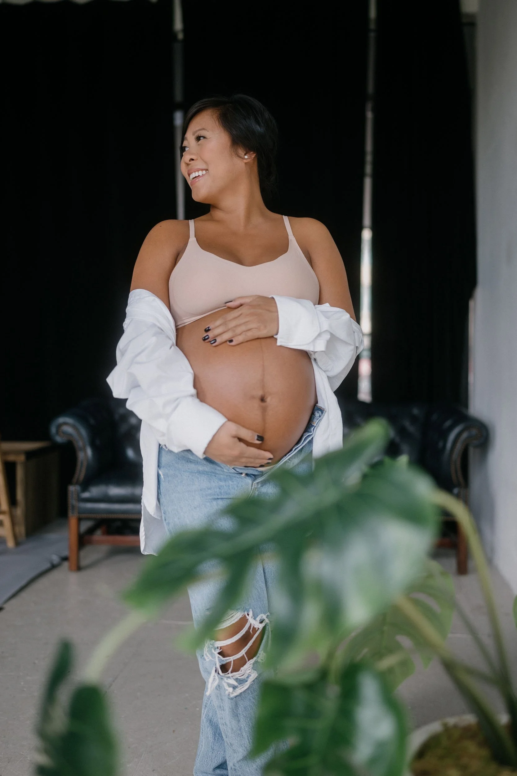 A pregnant woman smiling and looking to the side, standing indoors, wearing a beige top, ripped jeans, and an open white shirt, with black hair and dark nail polish.