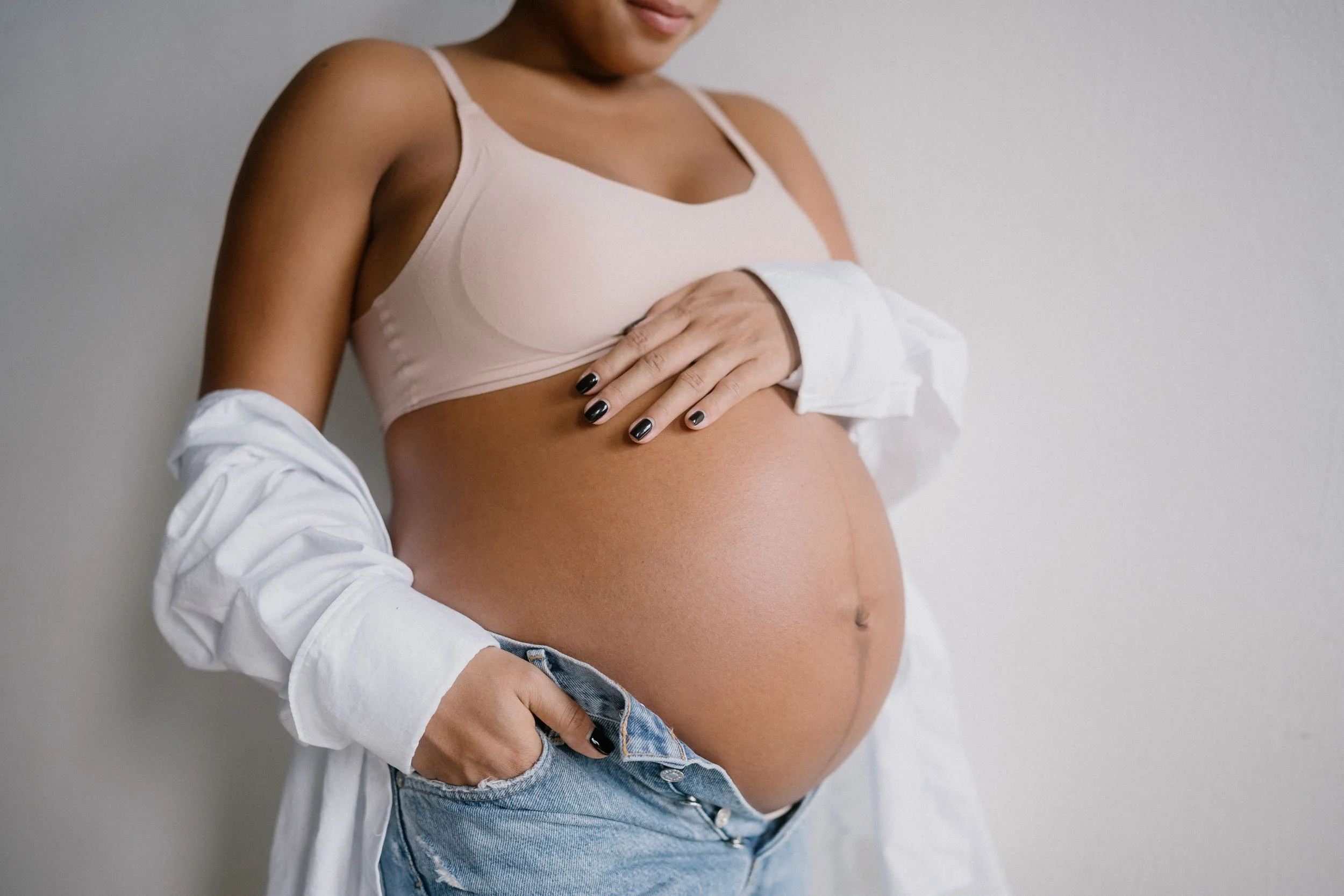 A pregnant woman with her hand on her belly, wearing a beige crop top and a white shirt, standing against a plain background.