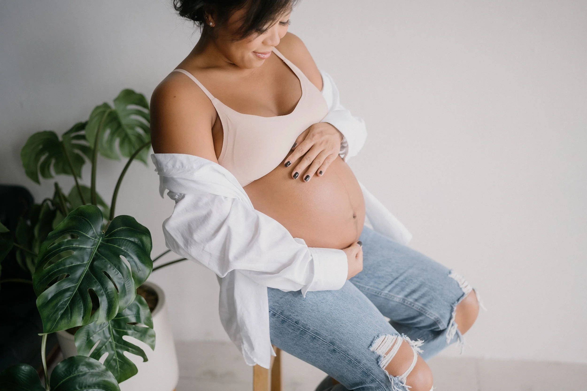 Pregnant woman sitting on a wooden chair, touching her belly, smiling, with large green houseplant nearby, in a bright room.