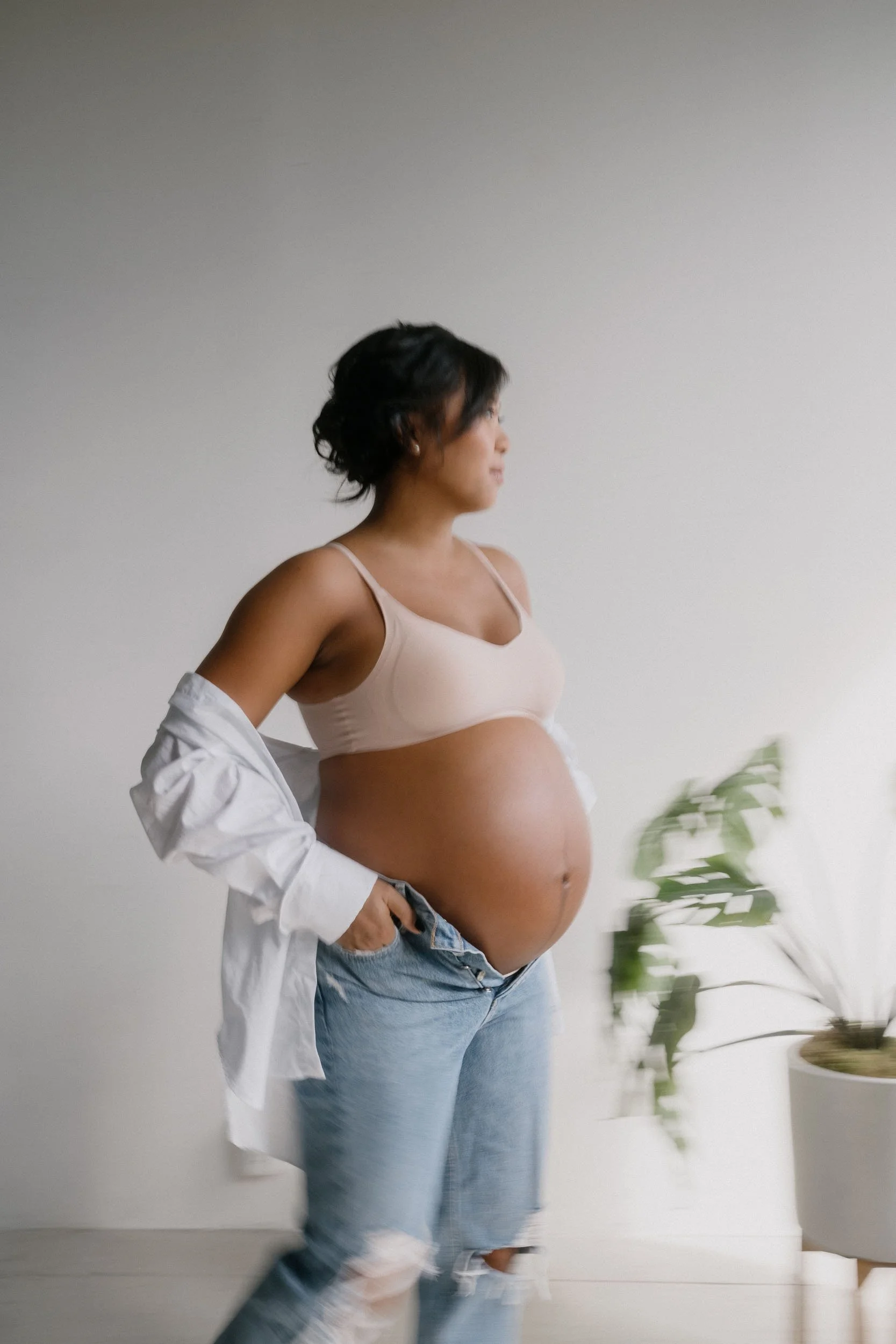 Pregnant woman standing indoors next to a potted plant, wearing a beige bra, ripped jeans, and an open white shirt.