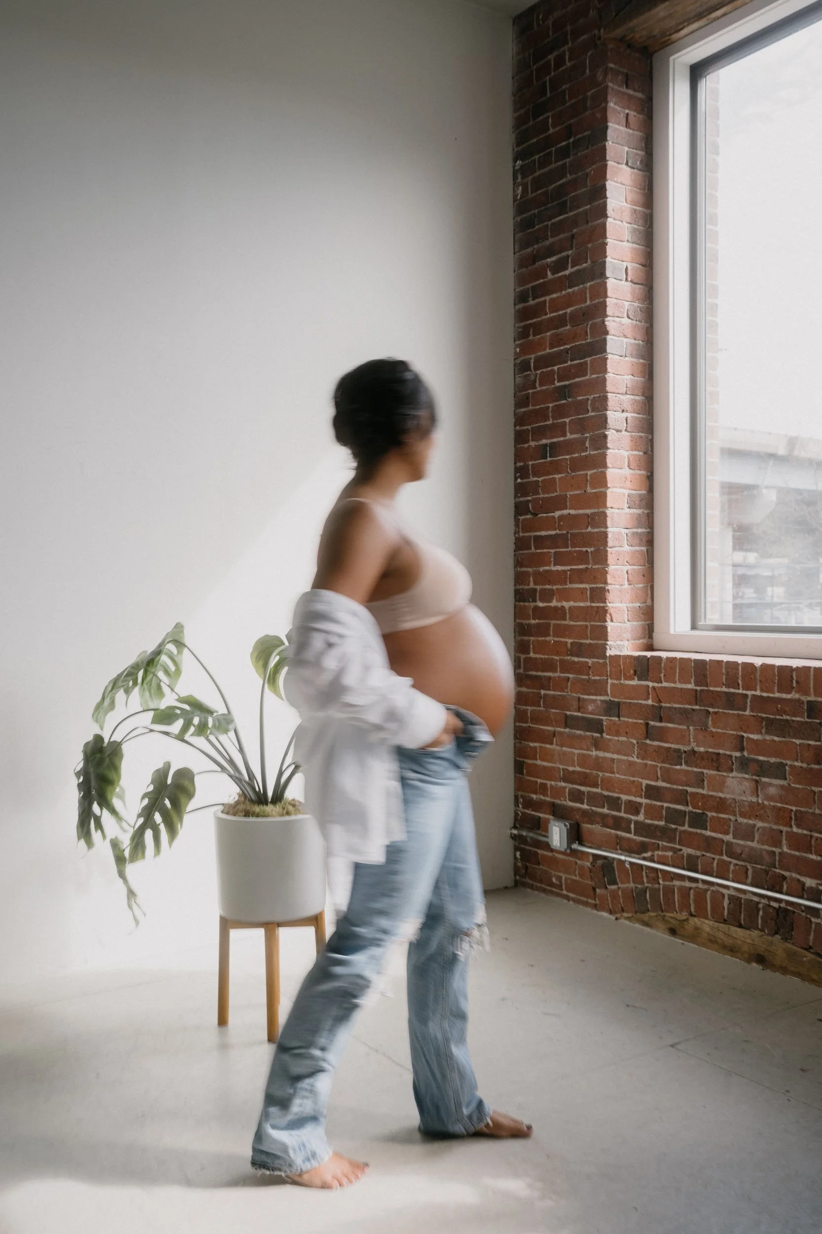 A pregnant woman in casual clothing standing in a minimalistic room with exposed brick wall, large window, and a potted plant.
