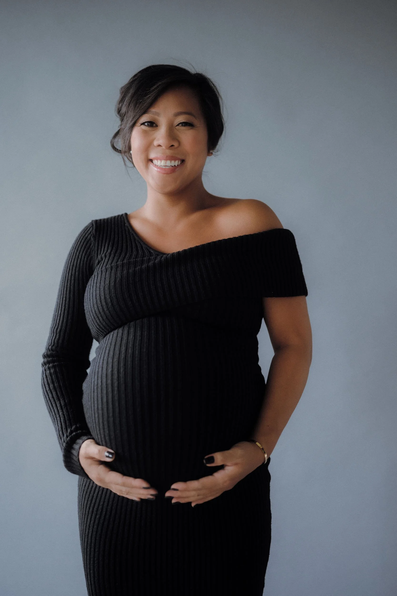 Portrait of a pregnant woman smiling, wearing a black off-the-shoulder ribbed dress, standing against a plain gray background.