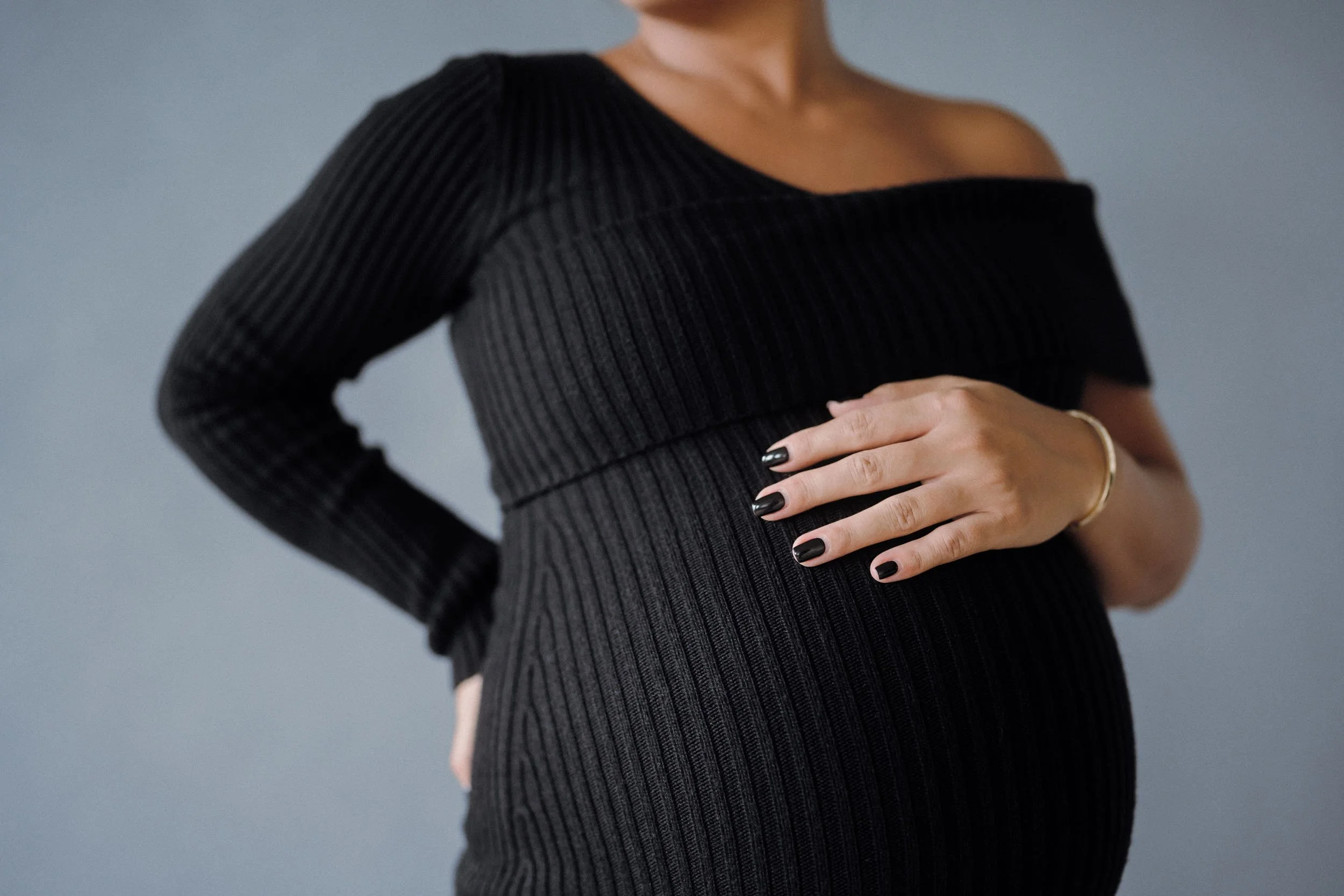 A woman in a black, ribbed, off-the-shoulder dress, holding her pregnant belly with one hand, wearing black nail polish and a gold bracelet, against a plain gray background.
