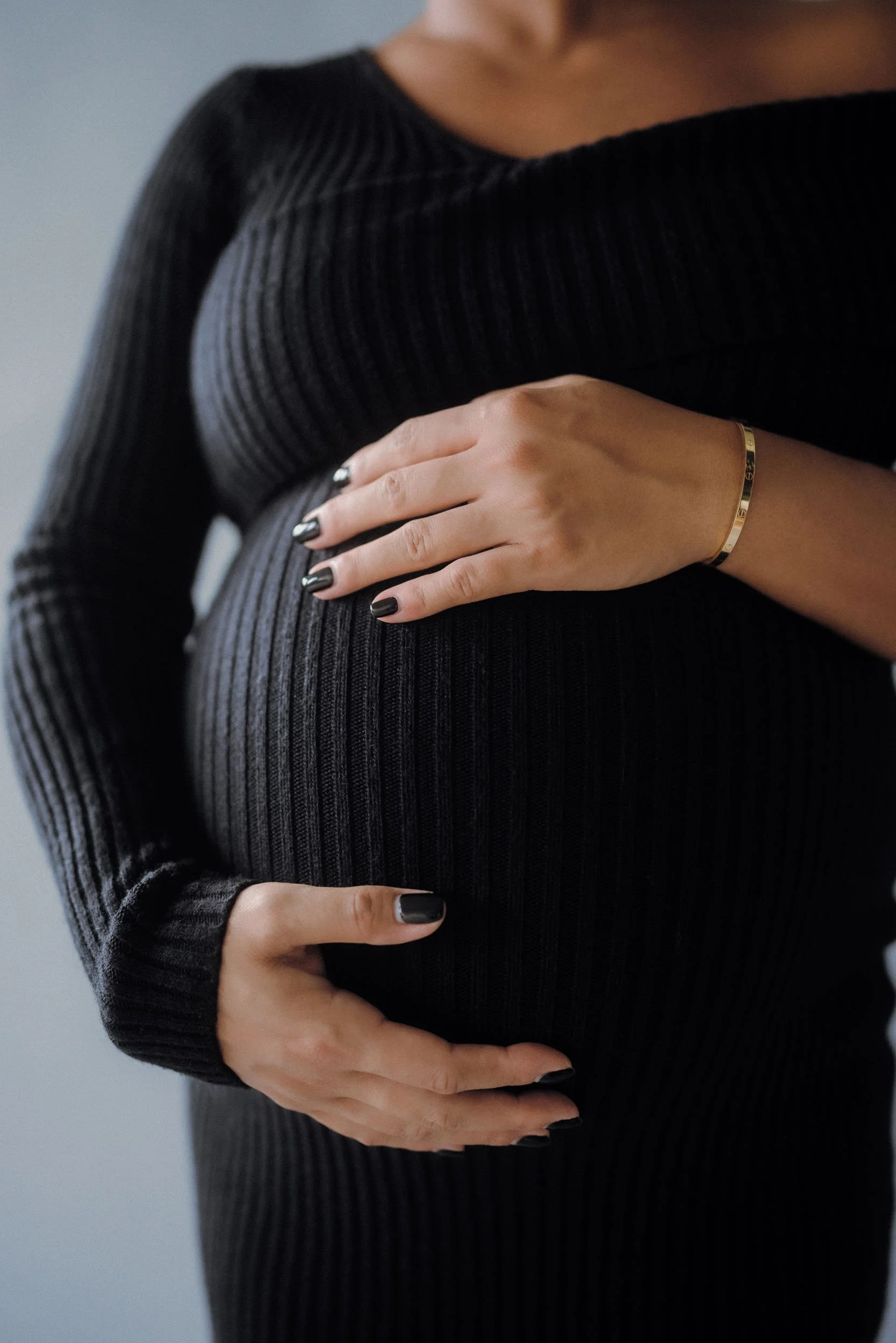 A pregnant woman in a black ribbed long-sleeve dress, holding her belly with one hand and the other hand resting on top, wearing a gold bracelet.
