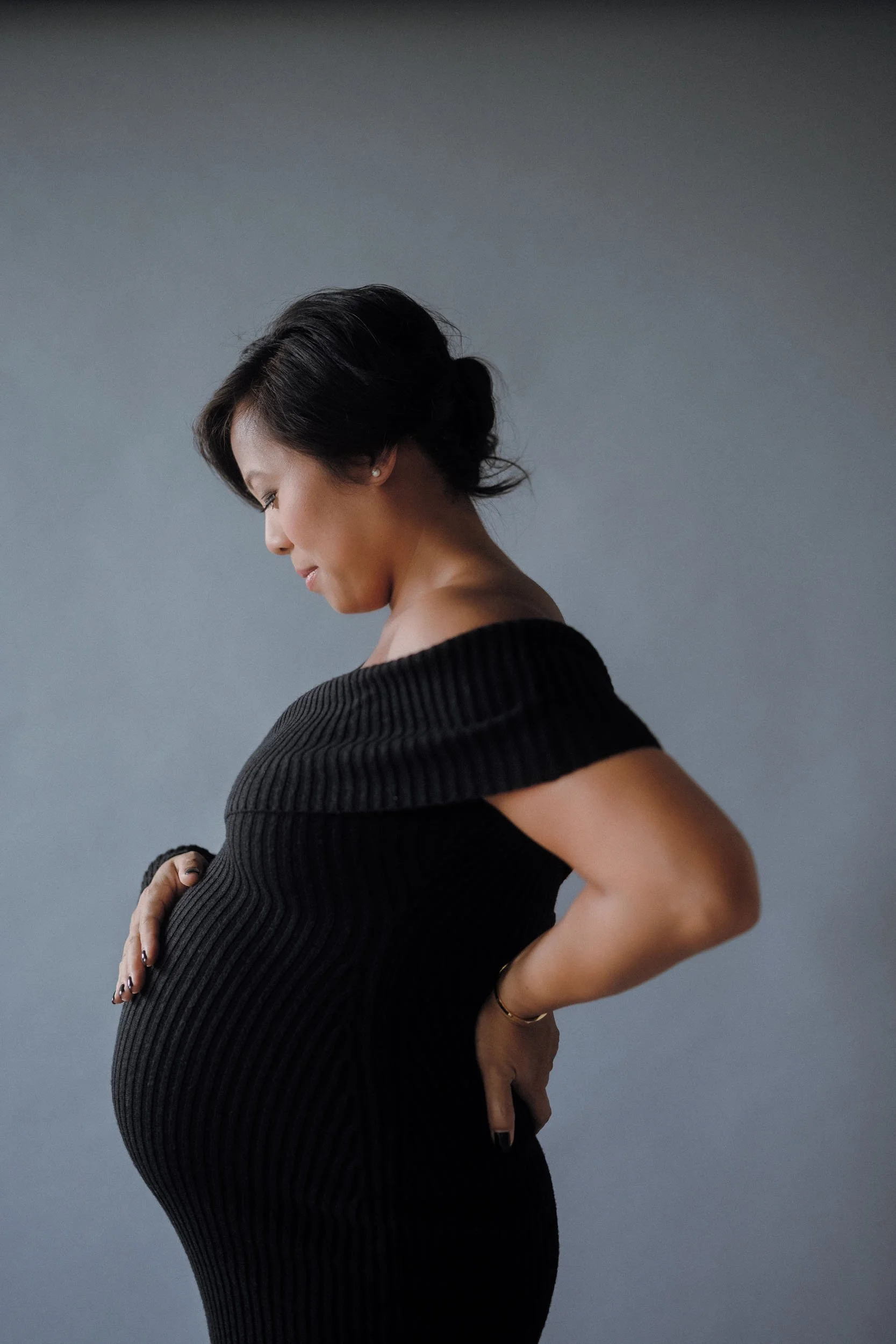Side view of a pregnant woman in a black ribbed off-the-shoulder dress, gently holding her belly, smiling softly, with short dark hair, against a plain gray background.