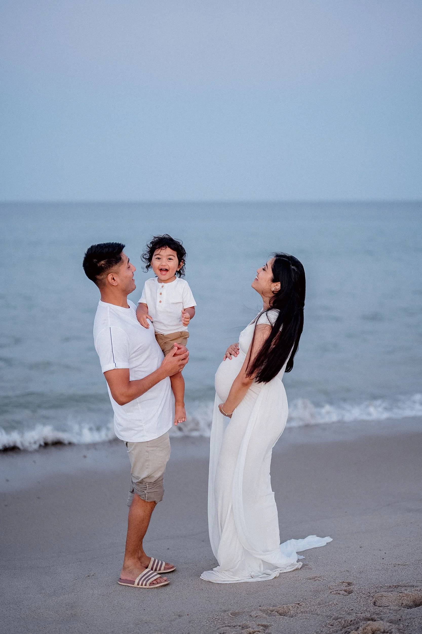 A happy family of three, including a pregnant woman, a man, and a girl, standing on the beach by the ocean during dusk, sharing joyful moments.