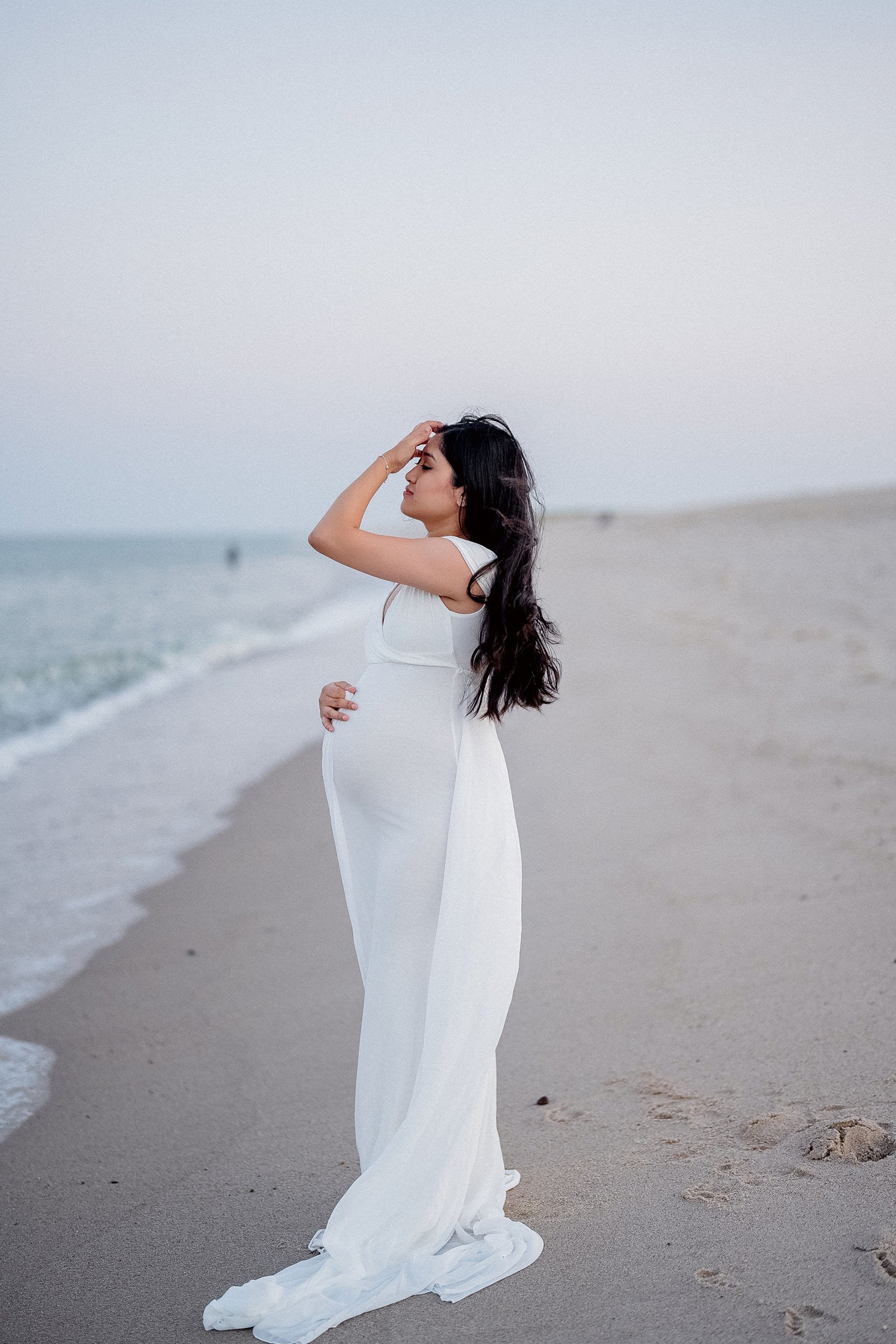 A pregnant woman in a white dress standing on the beach with her hand on her belly and her other hand on her head, near the water's edge.