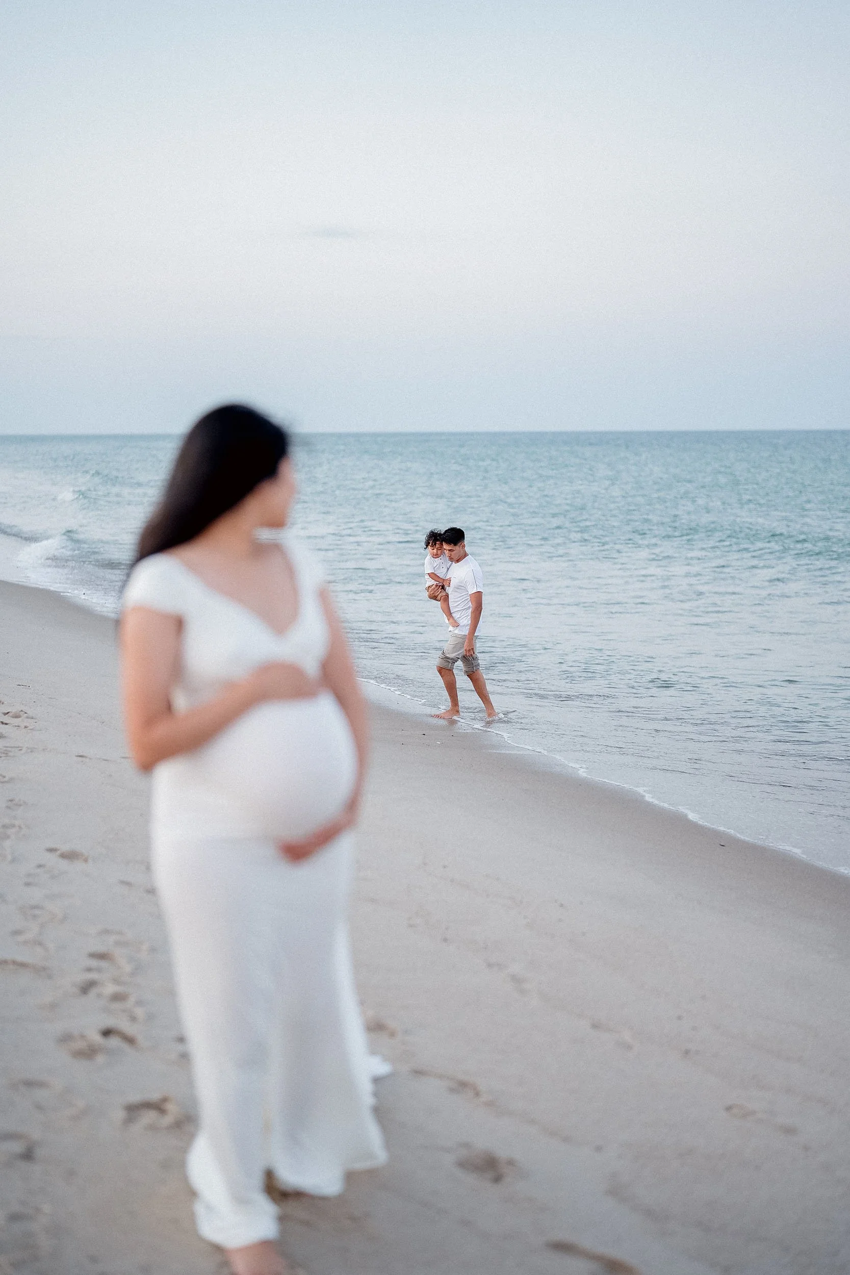 A pregnant woman in a white dress standing on the beach, with a man carrying a child in the background near the shoreline.