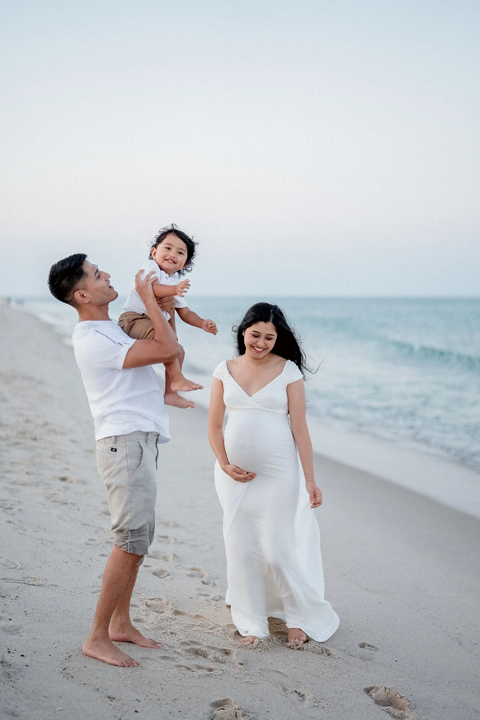 Family walking barefoot on the beach, with a woman pregnant, a man lifting a young girl, all smiling.