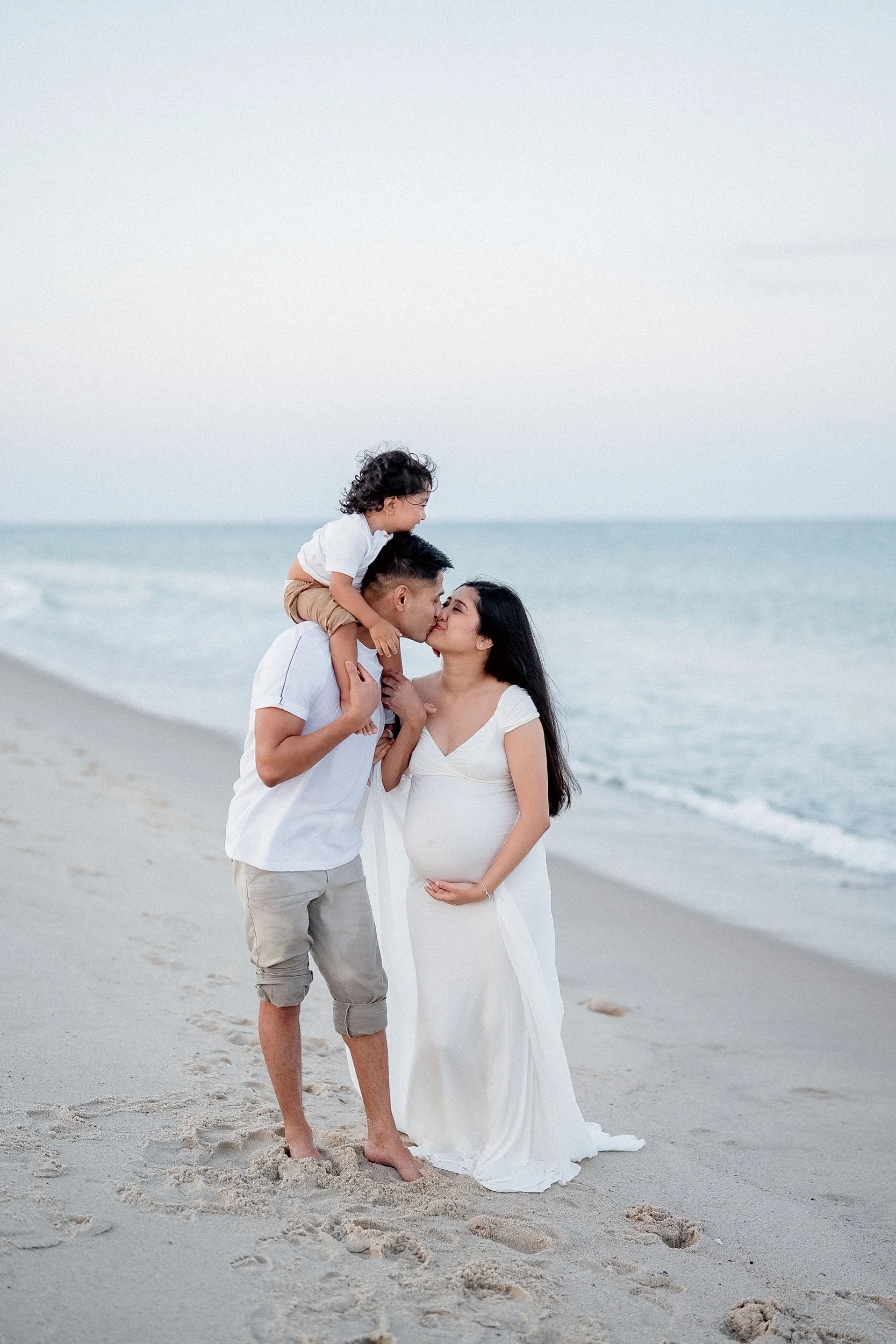 A family of three, including a pregnant woman, a man, and a young boy, sharing a kiss on the beach with the ocean in the background.