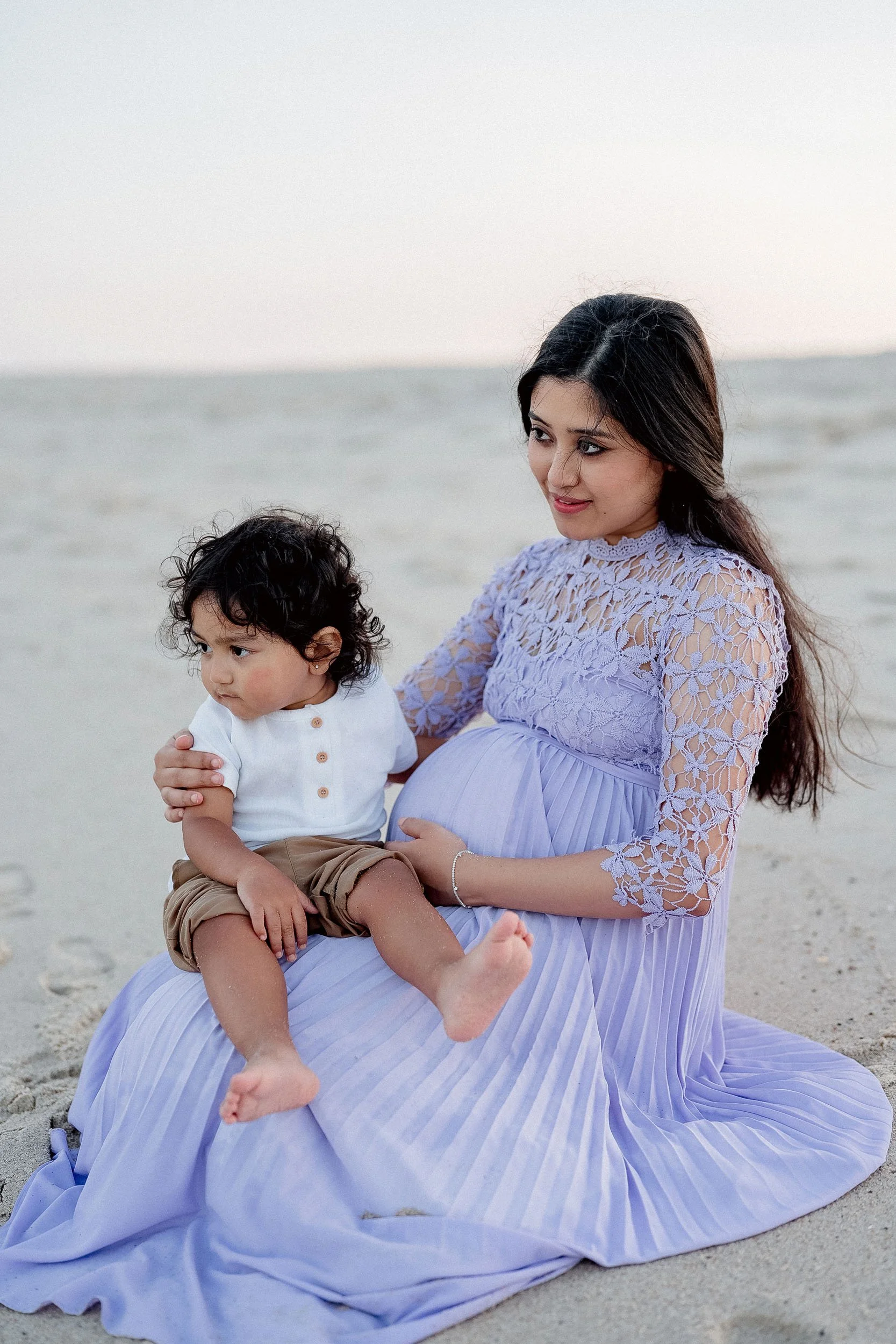 A pregnant woman sitting on the beach holding a young child in her lap, both looking thoughtful.