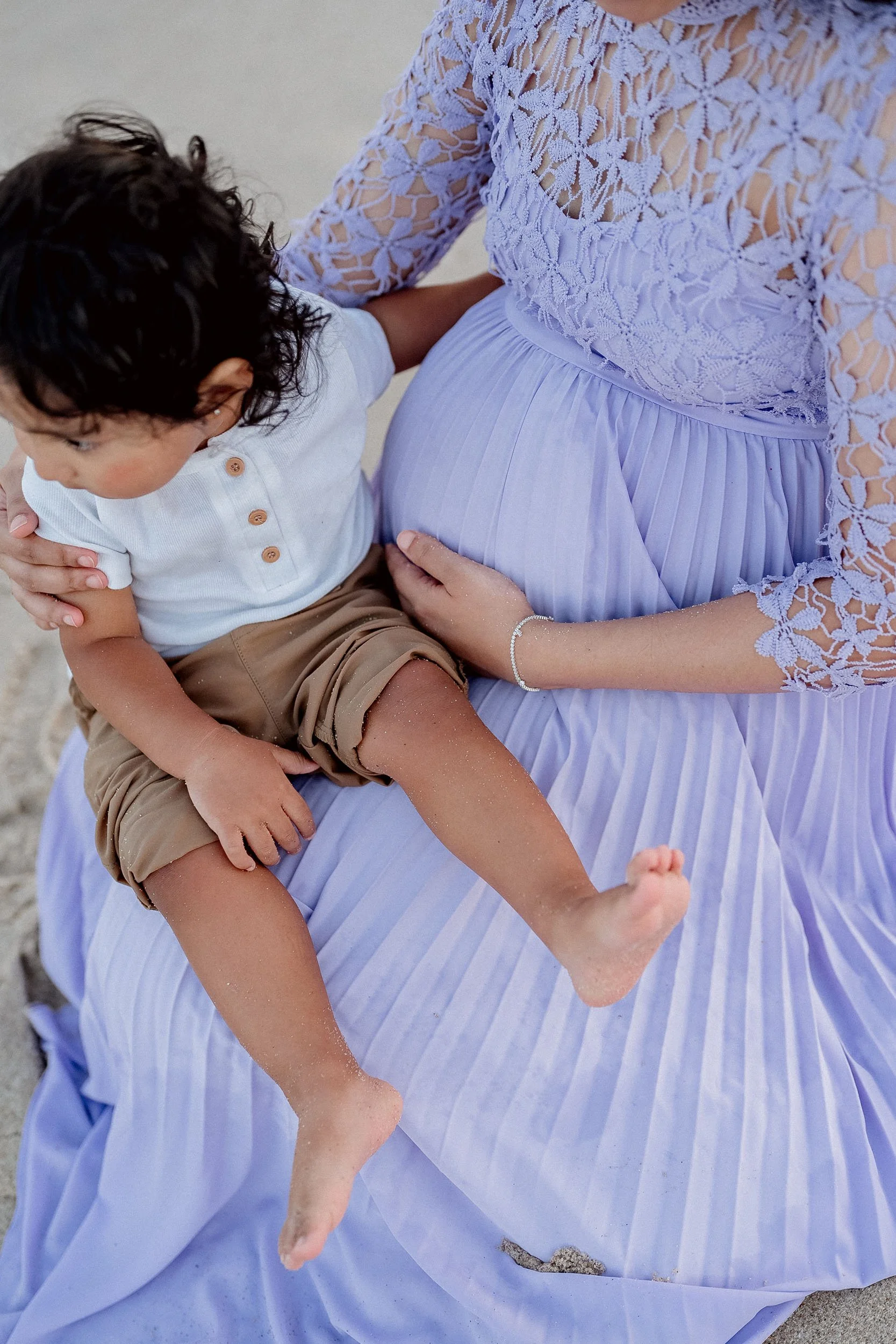 A pregnant woman in a lilac lace and pleated dress sitting on the sand, gently holding her belly with one hand, while a small child with dark curly hair in a white shirt and tan shorts sits on her lap, touching her belly.