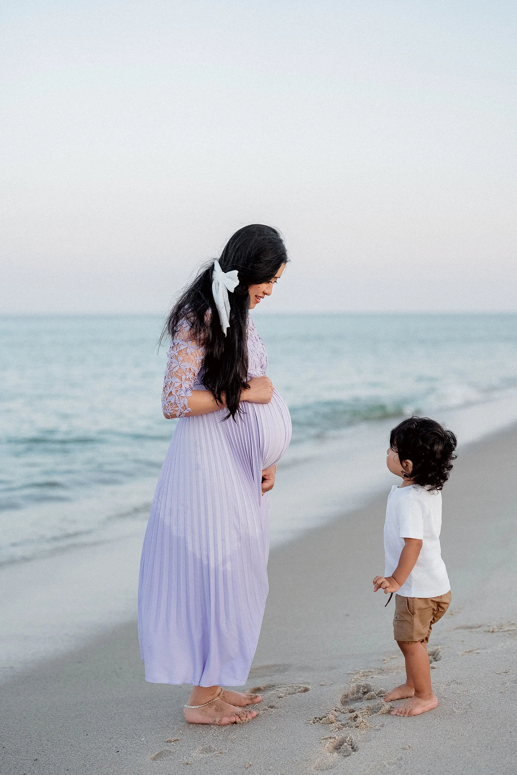 A woman and a young child standing barefoot on a beach, facing each other. The woman is pregnant, wearing a lavender dress with lace sleeves, and has a white bow in her hair. The child is wearing a white T-shirt and brown shorts, looking up at the wo