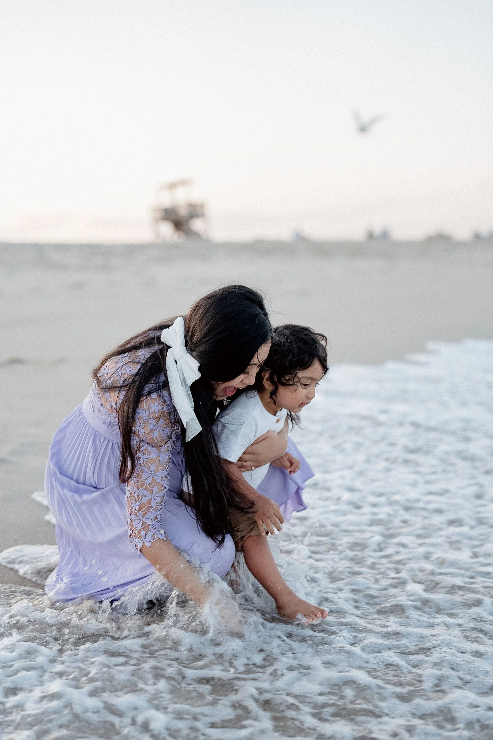 A woman and a young girl playing in the ocean water at the beach, with boats and a bird in the background.