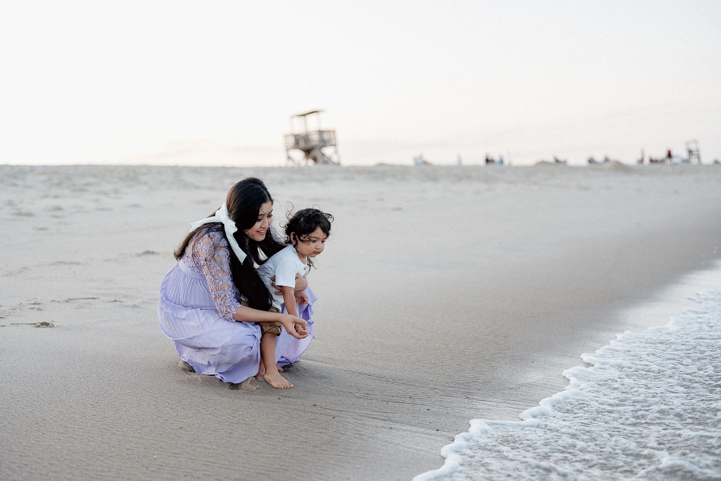 A woman and a young girl sitting on the sandy beach near the water's edge, looking at the sand.