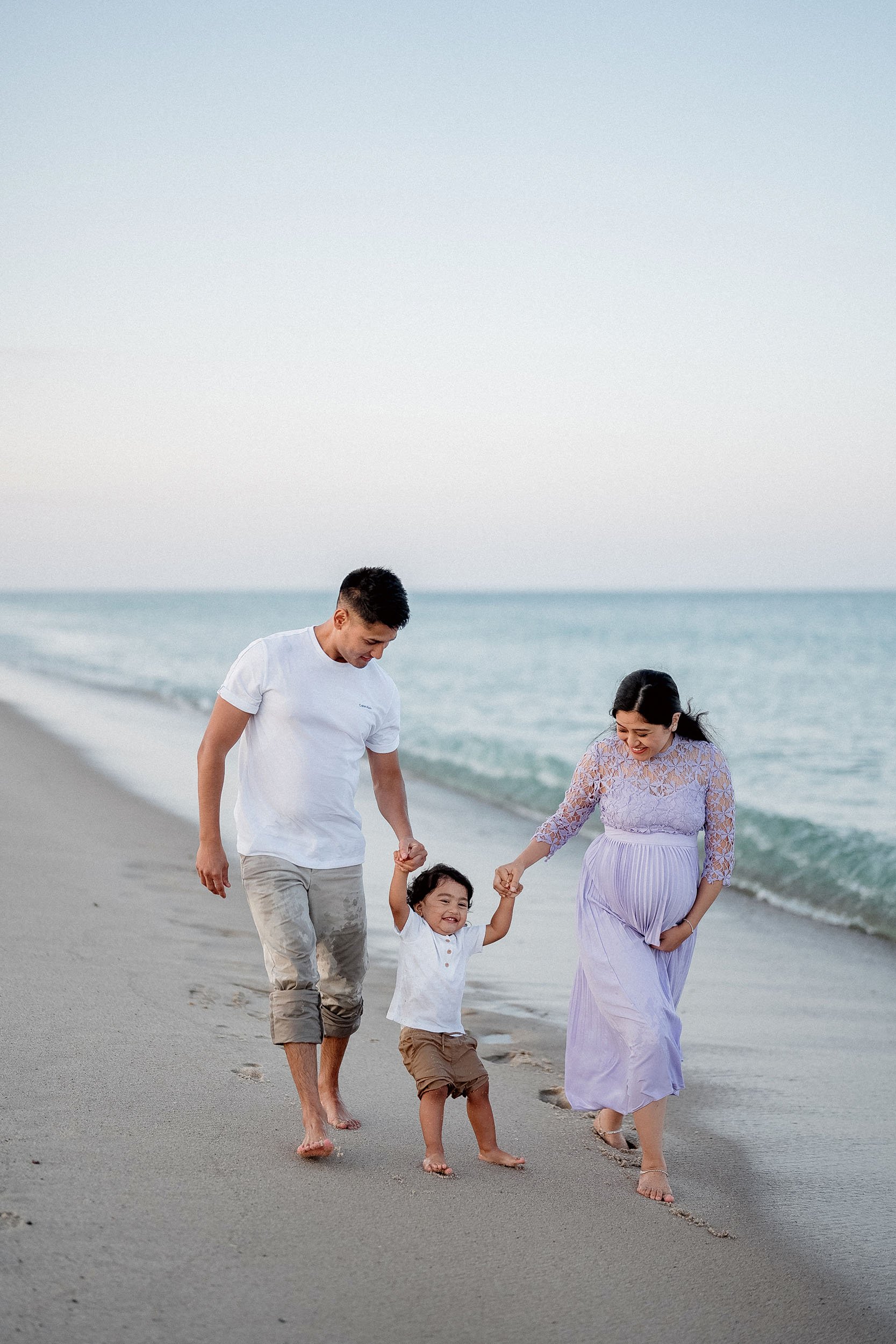 Family of three, a man, woman, and child, walking on the beach holding hands and smiling.