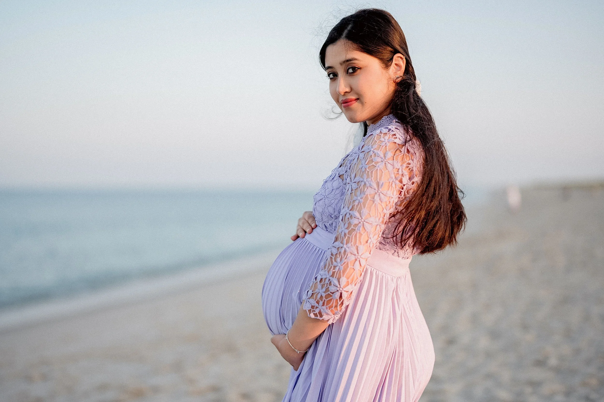 A pregnant woman with long dark hair standing on a beach, wearing a lavender dress with lace detail, looking at the camera with a gentle expression.