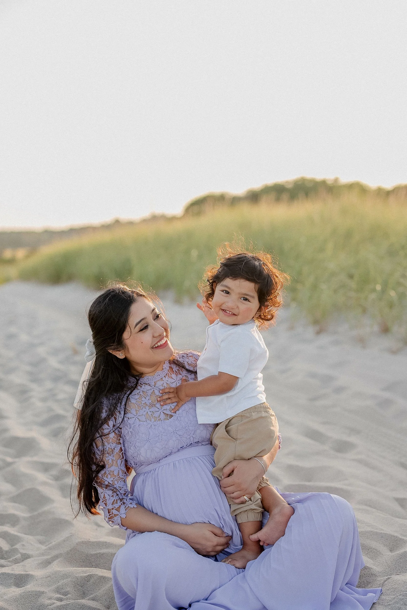 A woman and a young boy sitting on a sandy beach, smiling and playing together, with green dunes and a clear sky in the background during sunset.