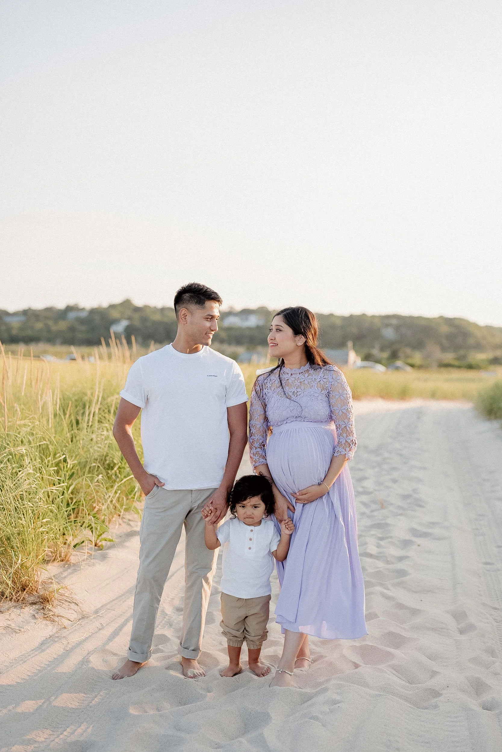 A pregnant woman, a man, and a young girl walk barefoot on a sandy beach with green grass on the sides during sunset. The woman is wearing a light purple dress with lace on the upper part, the man is in a white t-shirt and beige pants, and the girl i