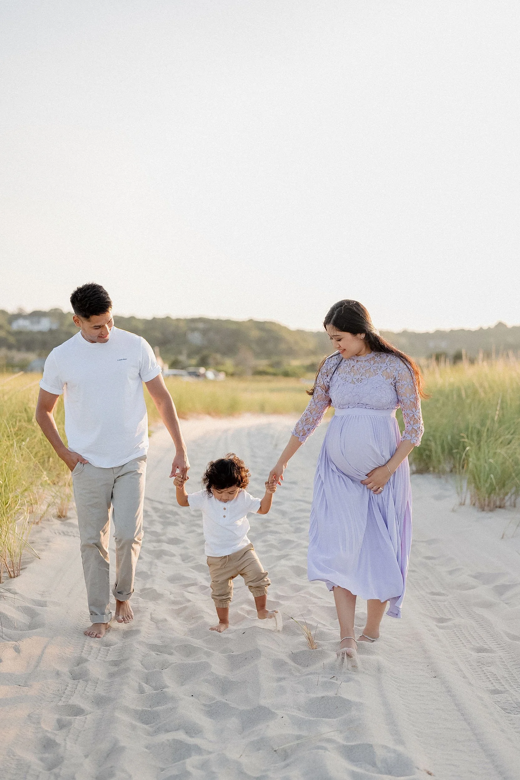 A family of three walking on a sandy beach, holding hands, with the mother visibly pregnant, during sunset.