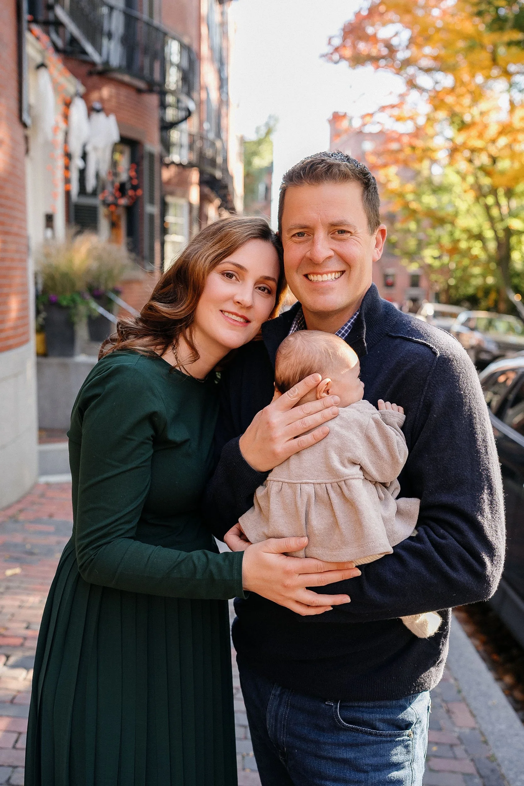 A happy family of three posing on a city street during autumn, with the mother, father, and their baby girl facing the camera and smiling, surrounded by fall foliage and brick buildings.