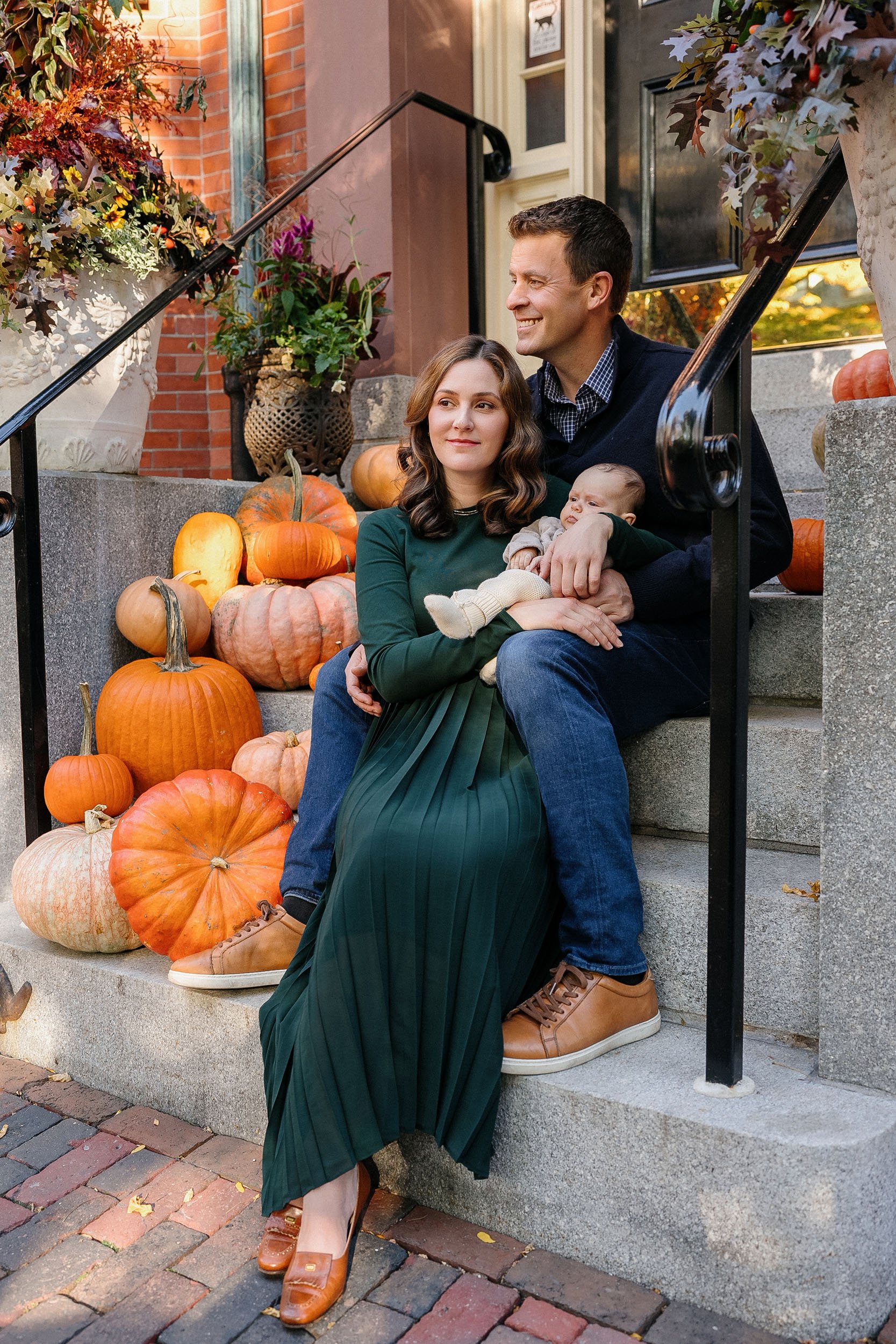 A family sitting on front steps decorated with pumpkins during fall. The man, woman, and baby are enjoying the crisp autumn weather on a porch with potted plants and a brick building in the background.