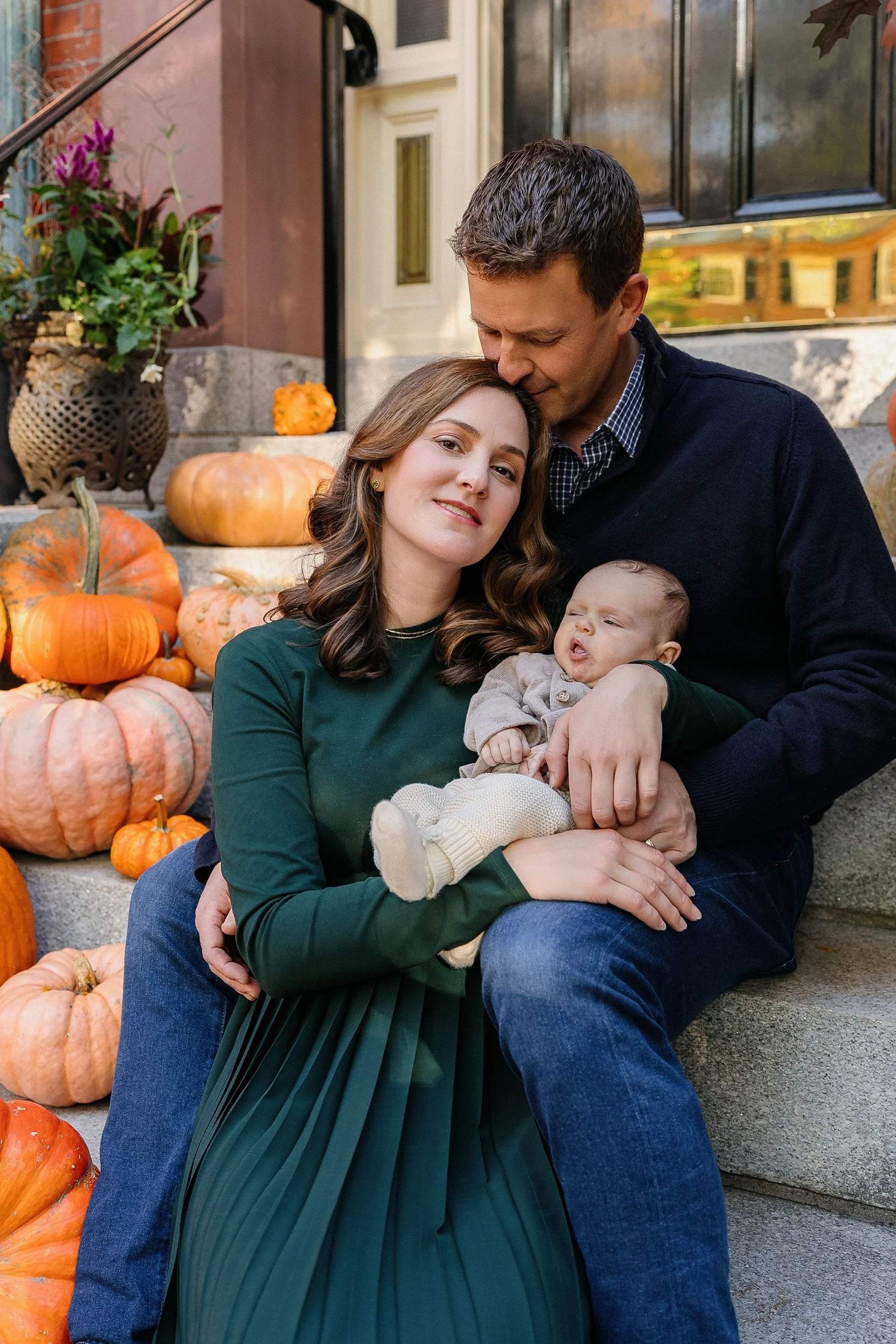 A family of three sitting on stairs decorated with pumpkins and autumn plants during fall. The mother has brown hair and is wearing a green dress, the father has short brown hair, and the baby is dressed in cozy clothing.