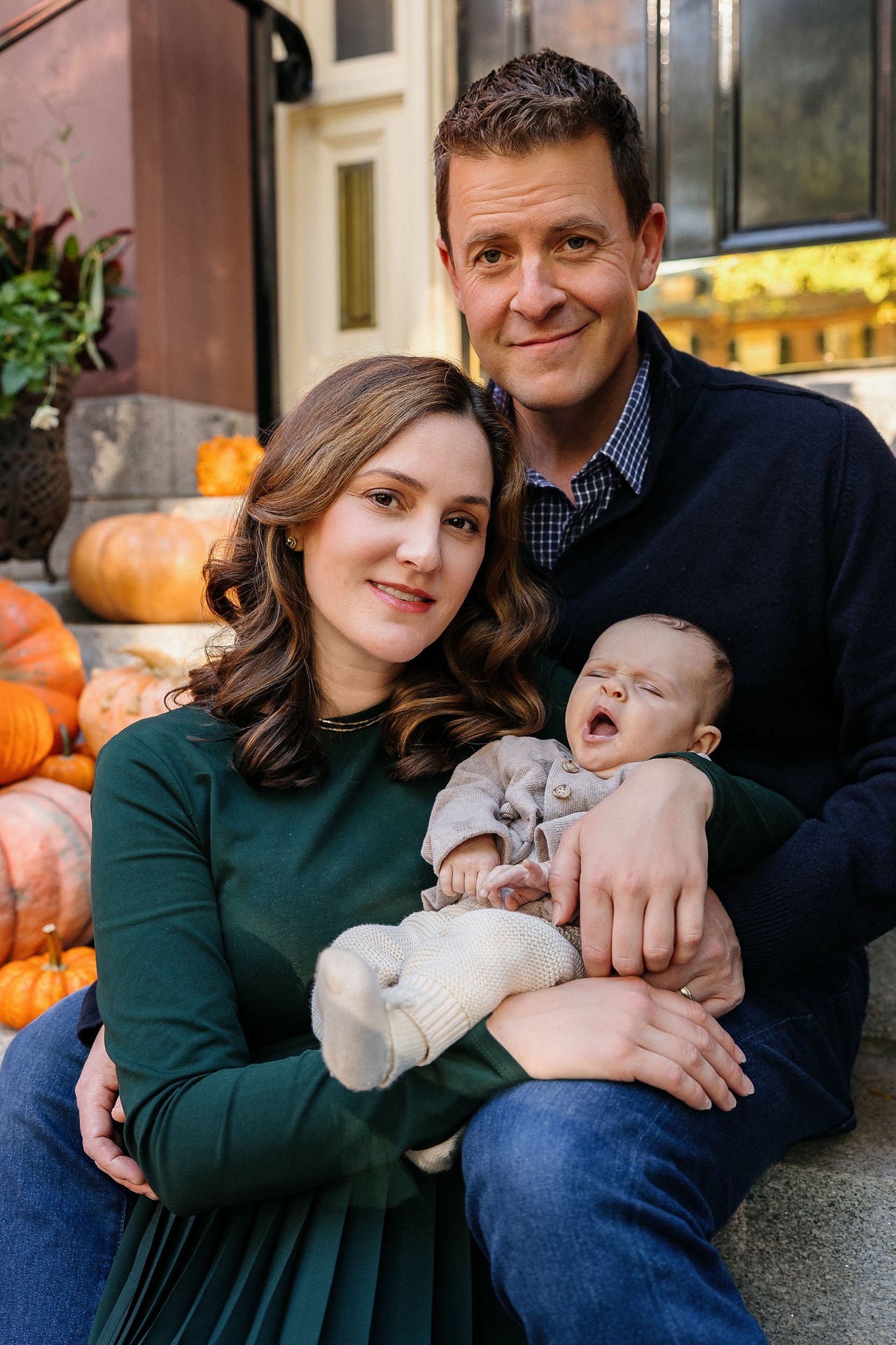 A family of three sitting on stairs outdoors, with autumn pumpkins around them. The mother, father, and a baby are posing for a photo, smiling at the camera.