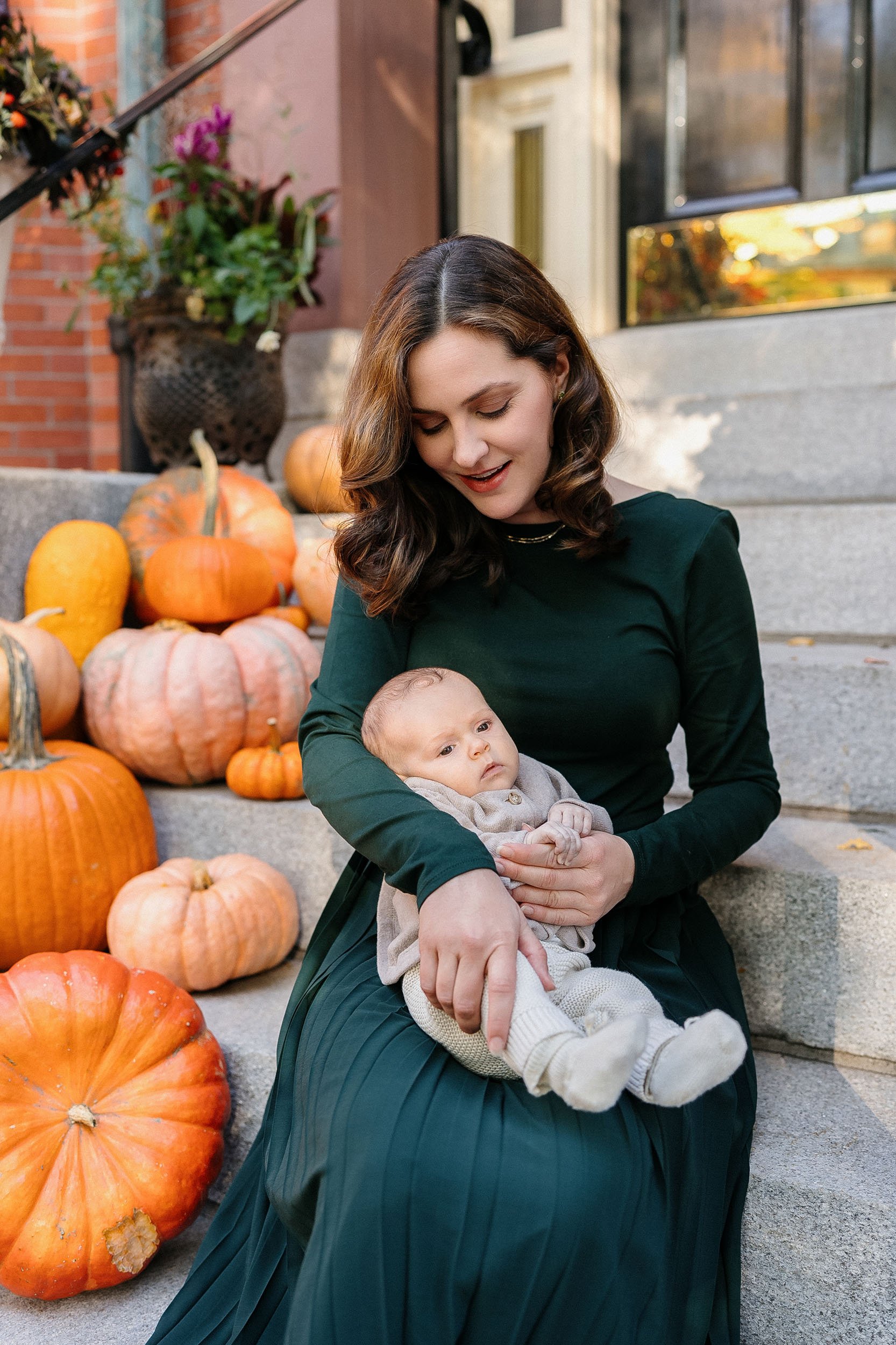 A woman sitting on steps surrounded by pumpkins, holding a baby in her lap, smiling and looking down at the baby