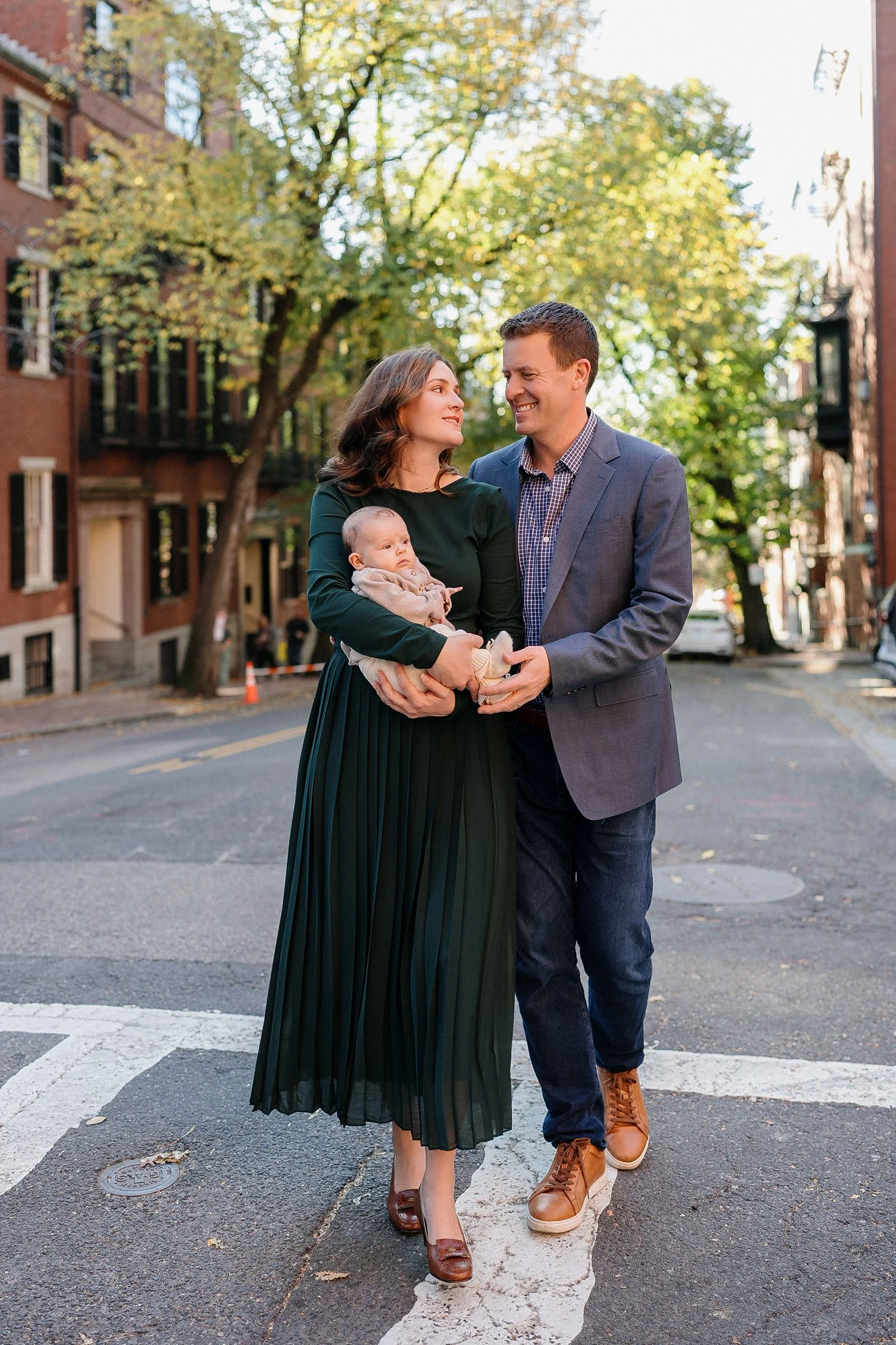 A family of three, a woman holding a baby and a man, walking on a city street with trees and buildings in the background.