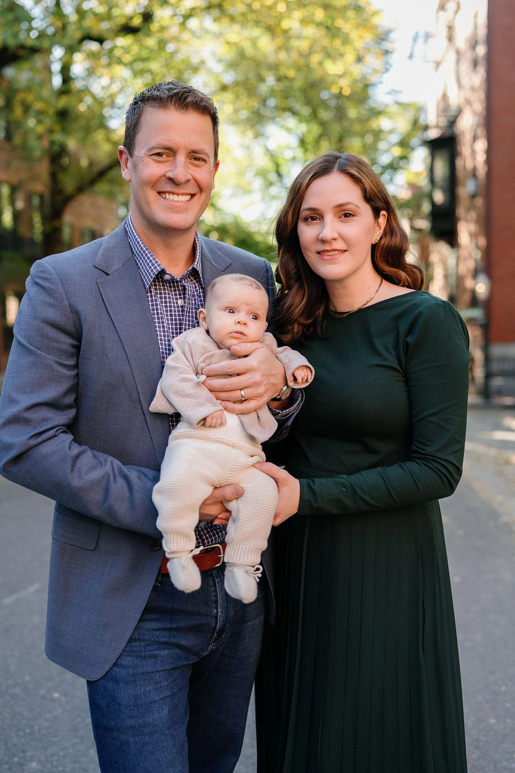 A family of three standing outdoors on a sunny day in a neighborhood with trees and buildings in the background. The man is holding a baby, and the woman is standing beside them.