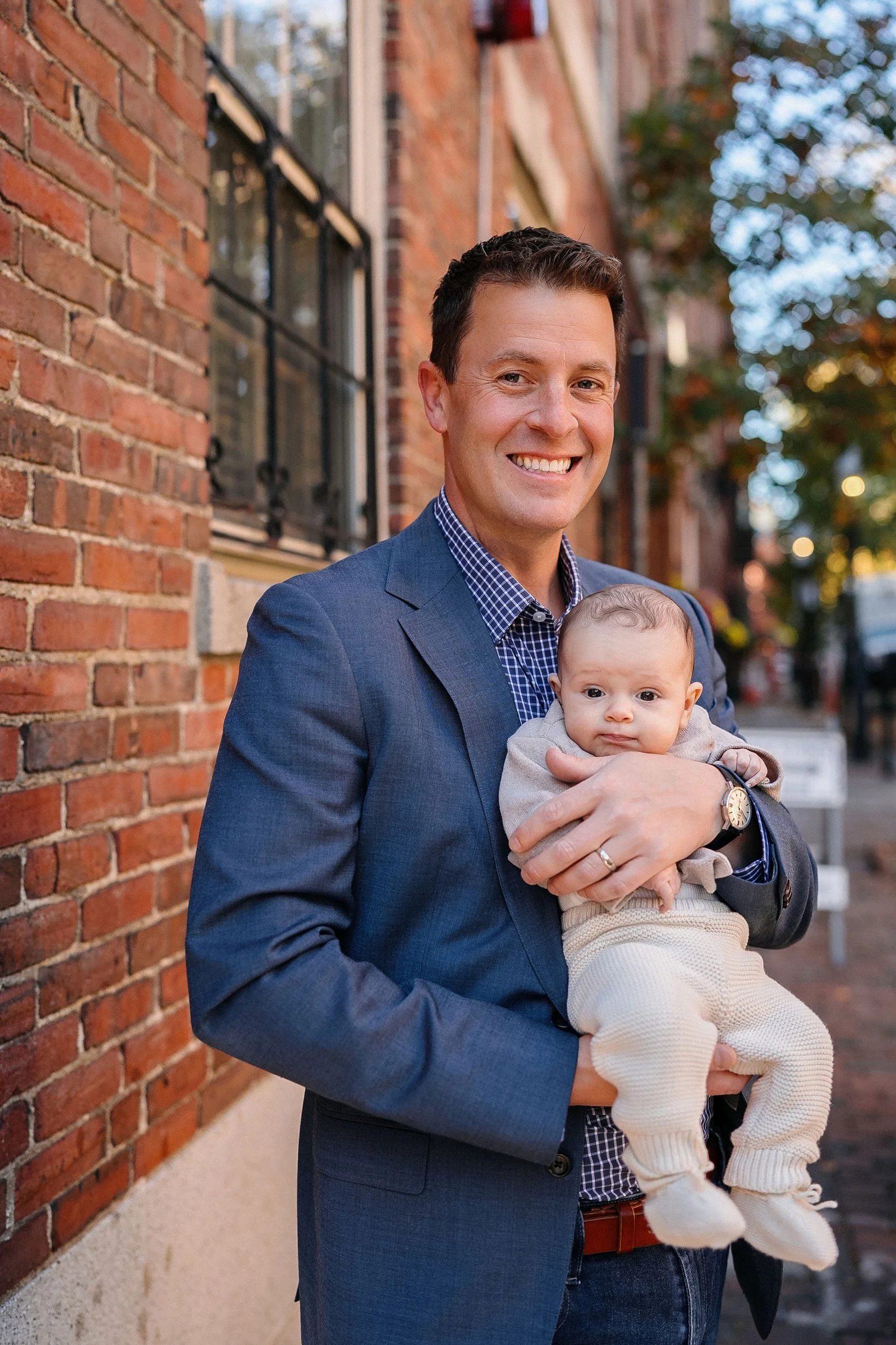A man in a blue suit smiling while holding a baby in his arms outside on a sidewalk in front of a brick building.