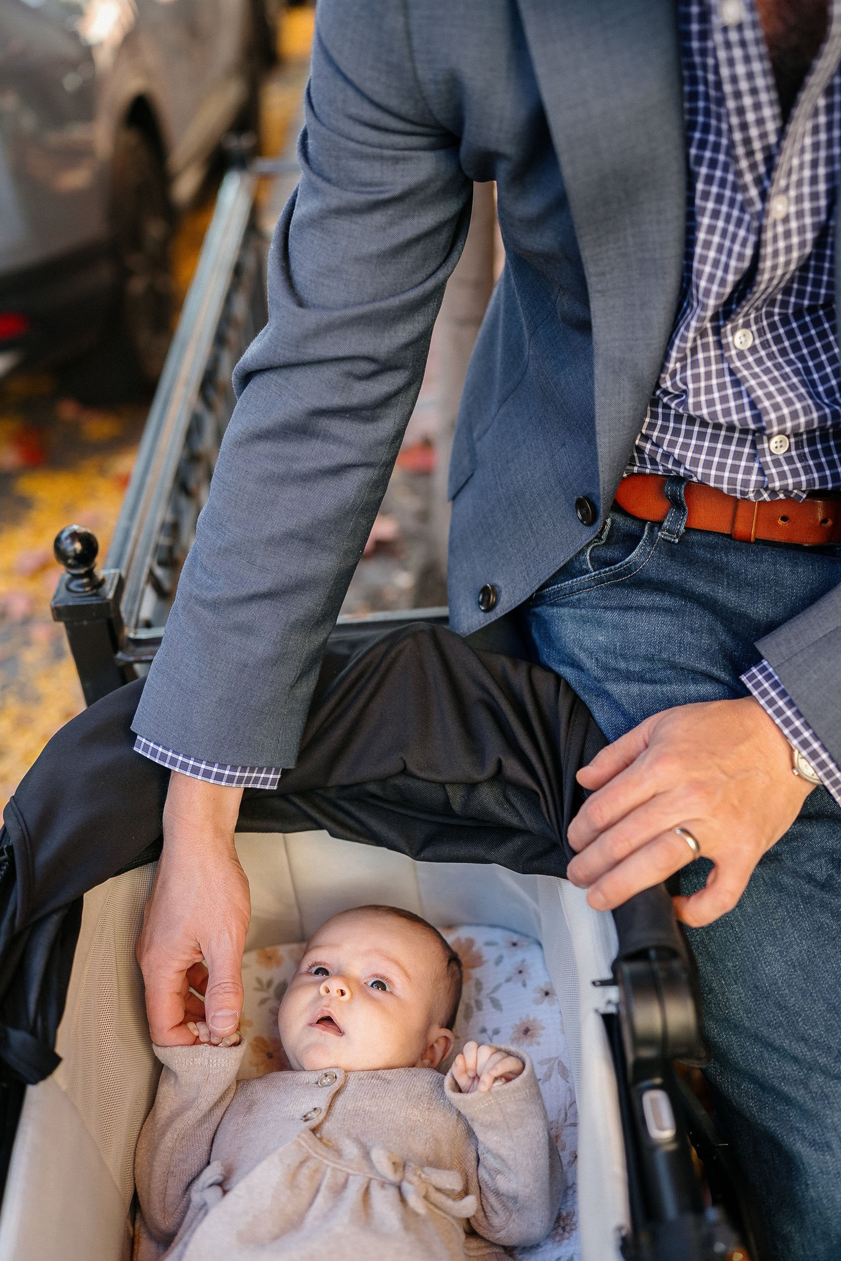 A man in a gray blazer and checkered shirt leaning over a stroller with a baby inside. The man is holding the stroller handle with one hand and the baby’s hand with the other. The baby is laying in the stroller, looking up with wide eyes, wearing a b